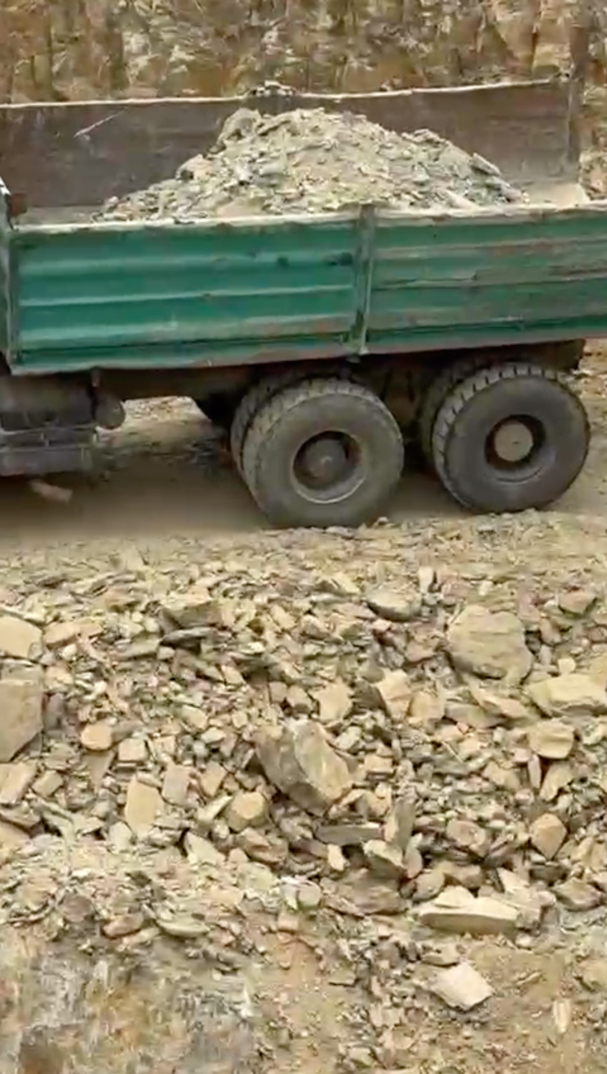 A video shows a rocky landscape. A green truck has it’s bed filled with grey rocks. Next to the truck is a yellow digger. The camera pans across and zooms in to show the rocky landscape.