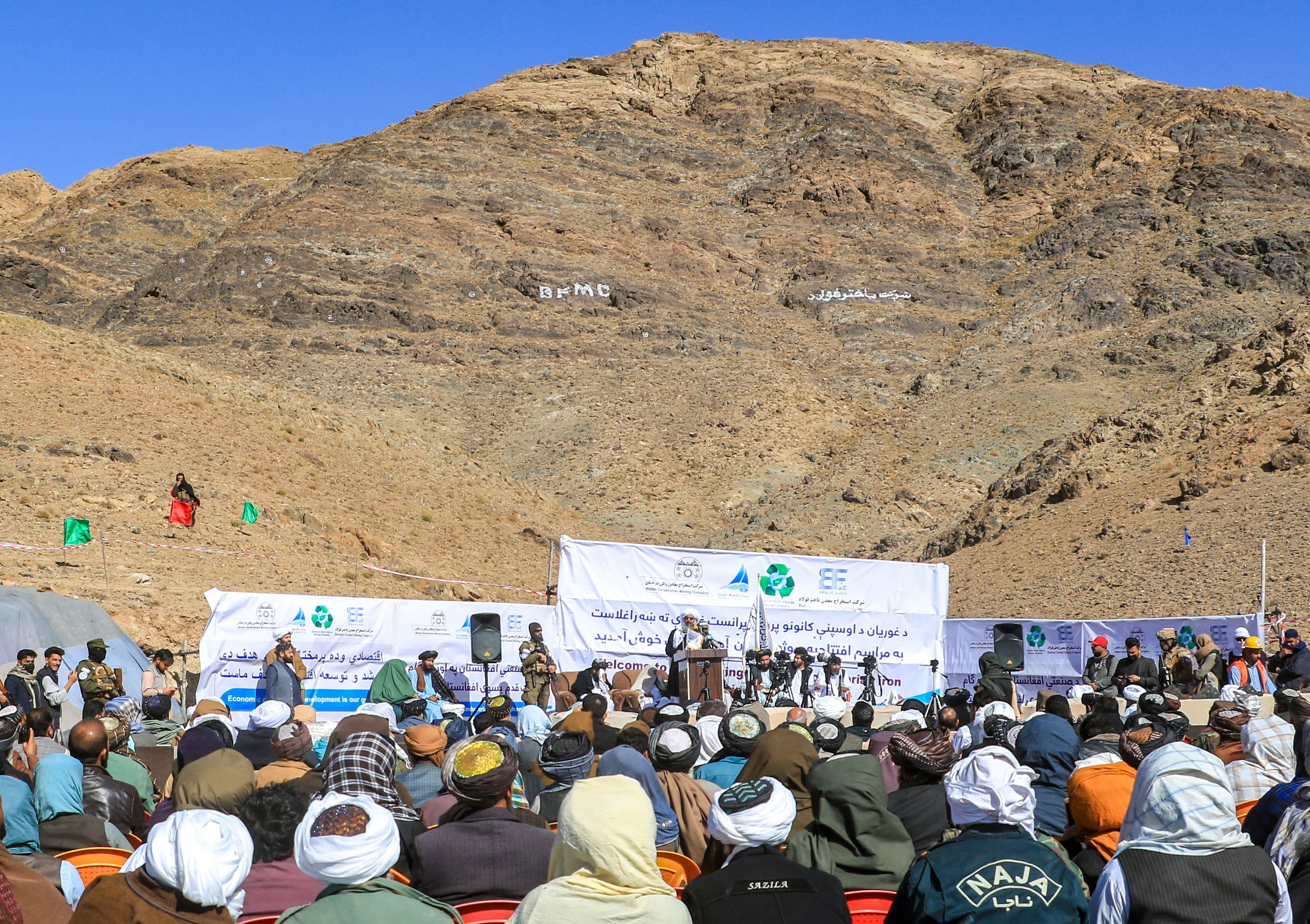 Image showing a crowd attending an event in front of a mountainous terrain. Men are seated before a lectern which has a speaker standing at it. Behind the speaker are posters.