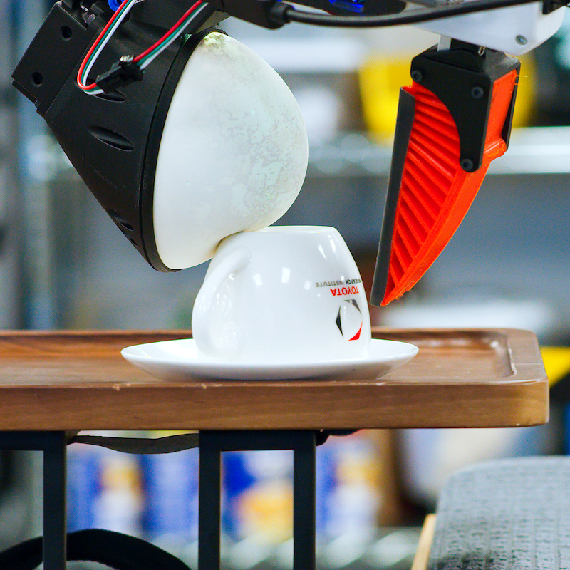 A robot arm with grippers attempts to pick up an upside down white teacup from a white saucer