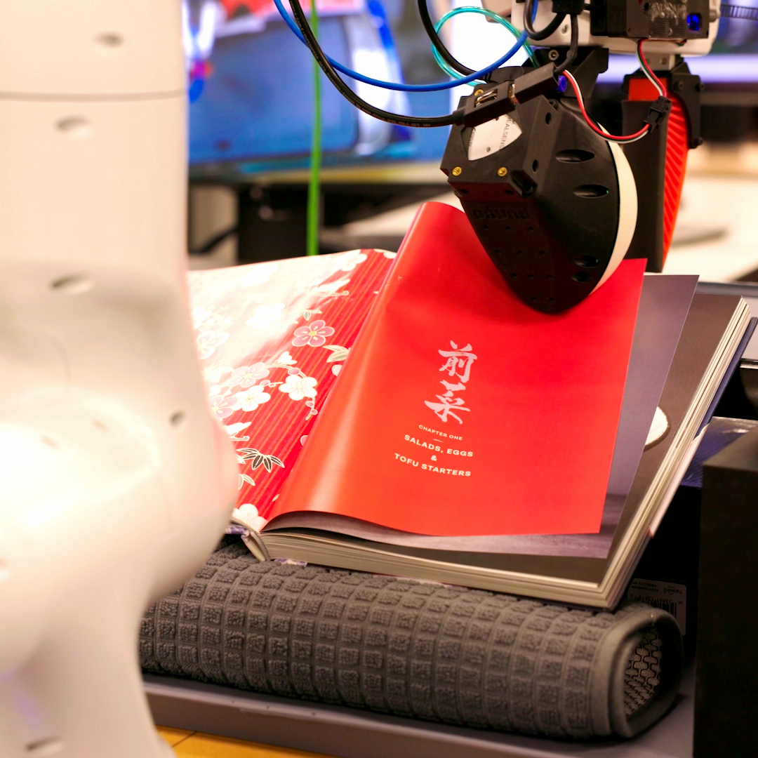 A white robot arm turns the pages of a book containing pictures of flowers
