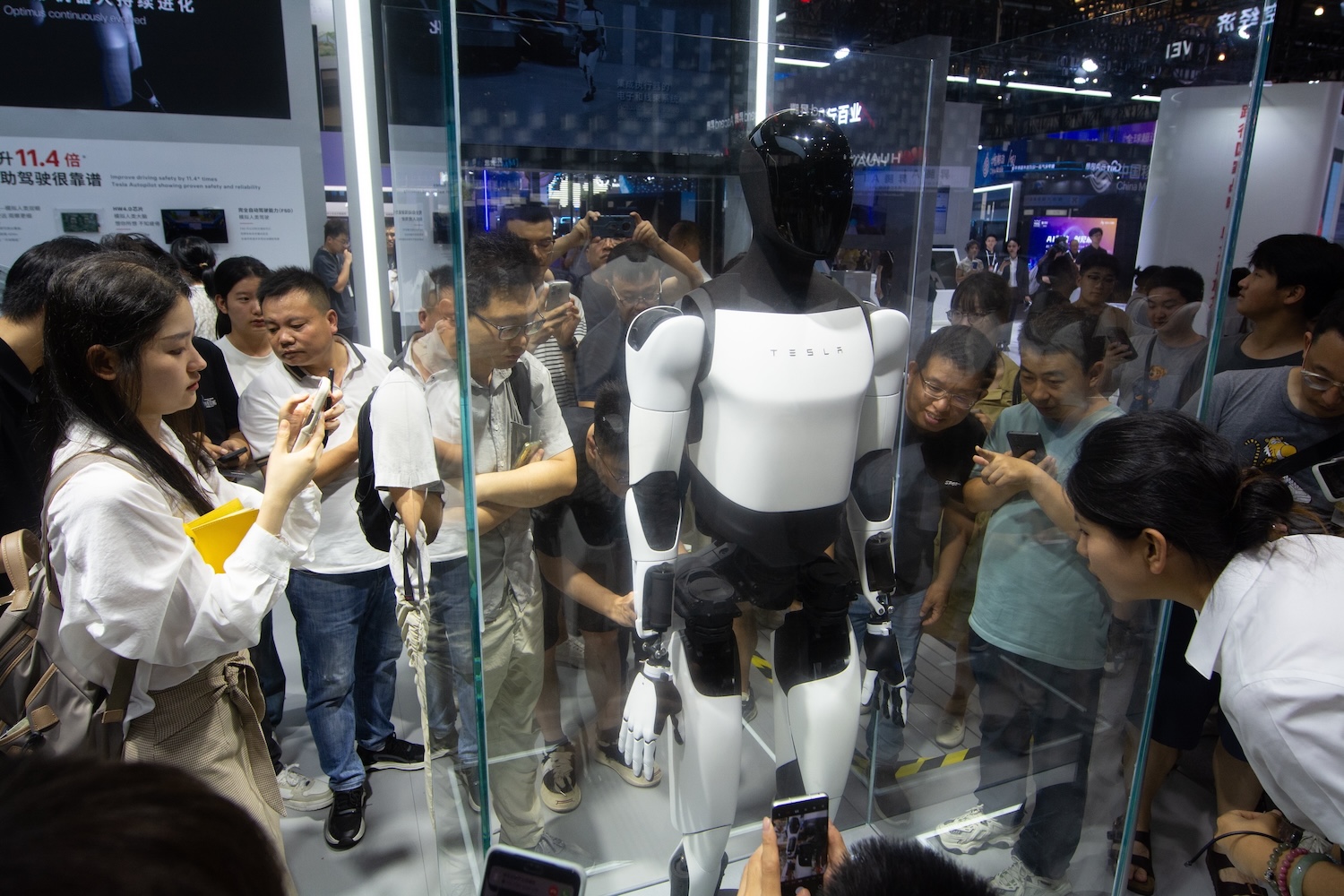 A crowd of people gather around a glass display case observing a Tesla Optimus humanoid robot with its sleek black-and-white design. Onlookers at the tech event appear curious.