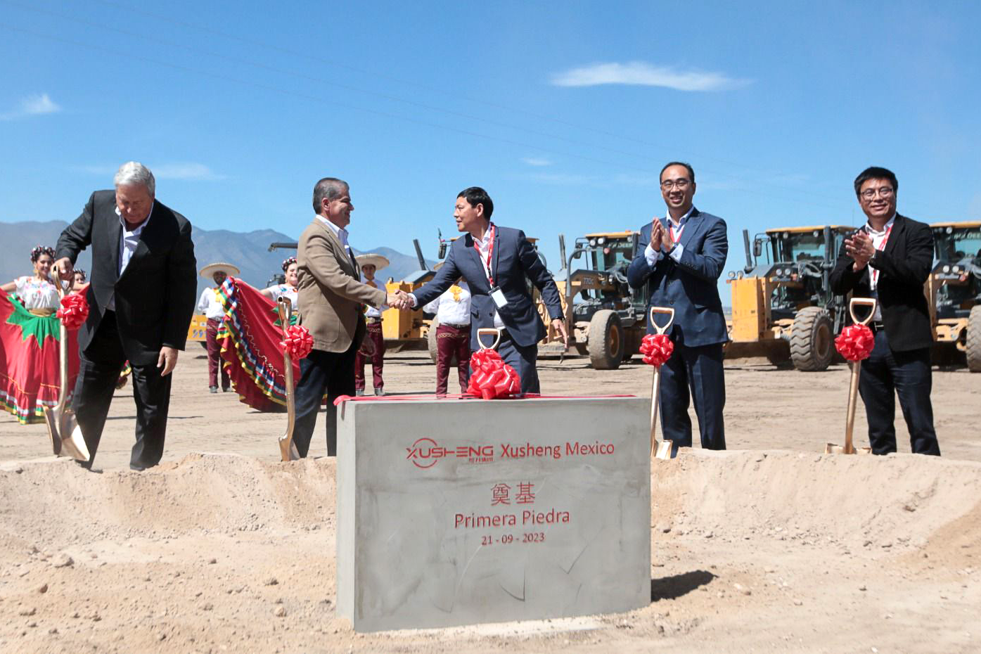 Local state officials and company executives in suits shake hands and applaud at a groundbreaking ceremony for Xusheng at Alianza Industrial Park in Coahuila state, Mexico, with construction equipment and dancers in colorful attire in the background.