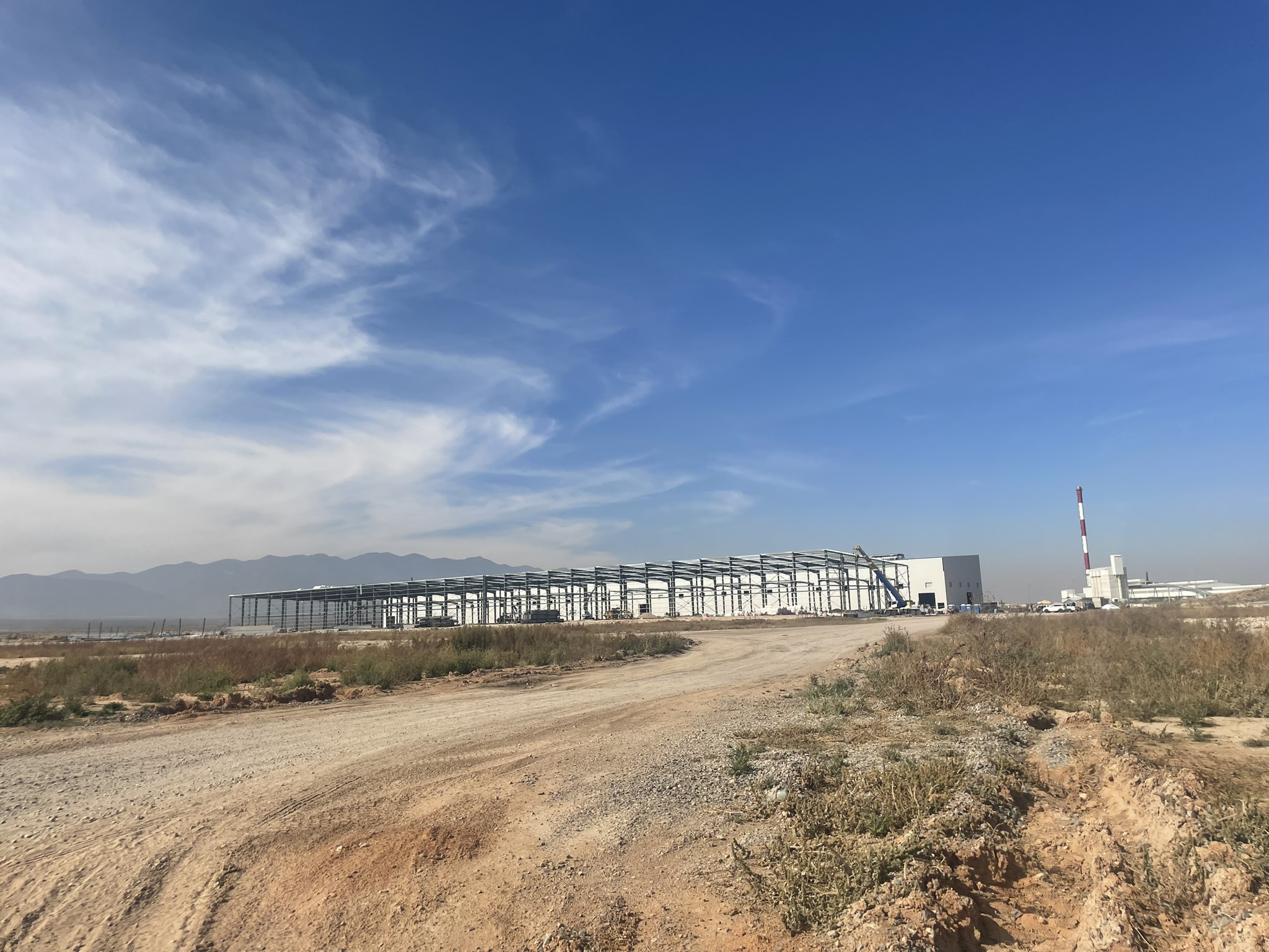 Image of a white industrial building under construction behind a grassy field with a clear blue sky and distant mountains in the background at Alianza Industrial Park in Coahuila state, Mexico