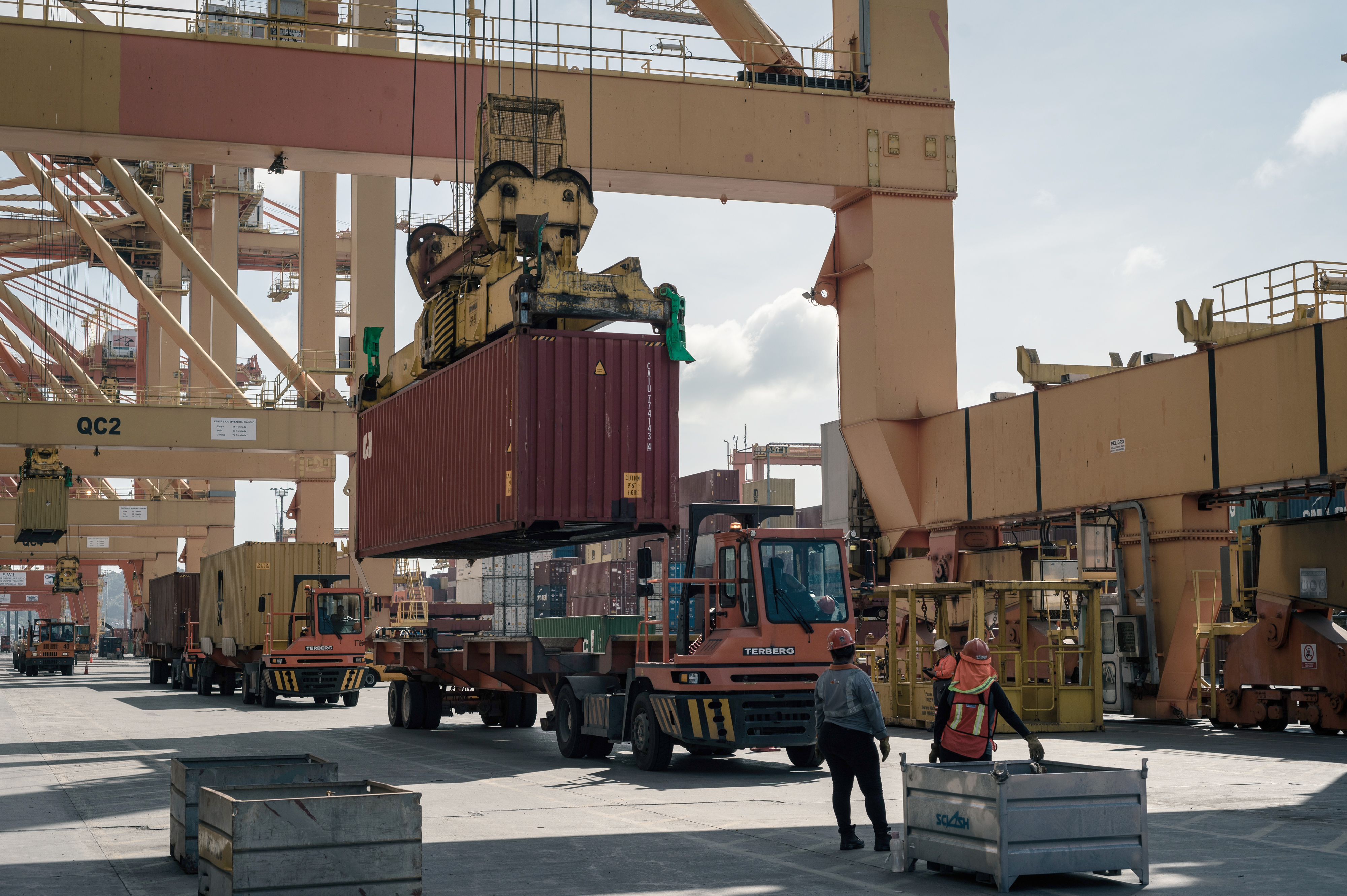 A gantry crane lifts a shipping container at the Port of Manzanillo, with port workers standing in the foreground and trucks with additional containers lining up behind