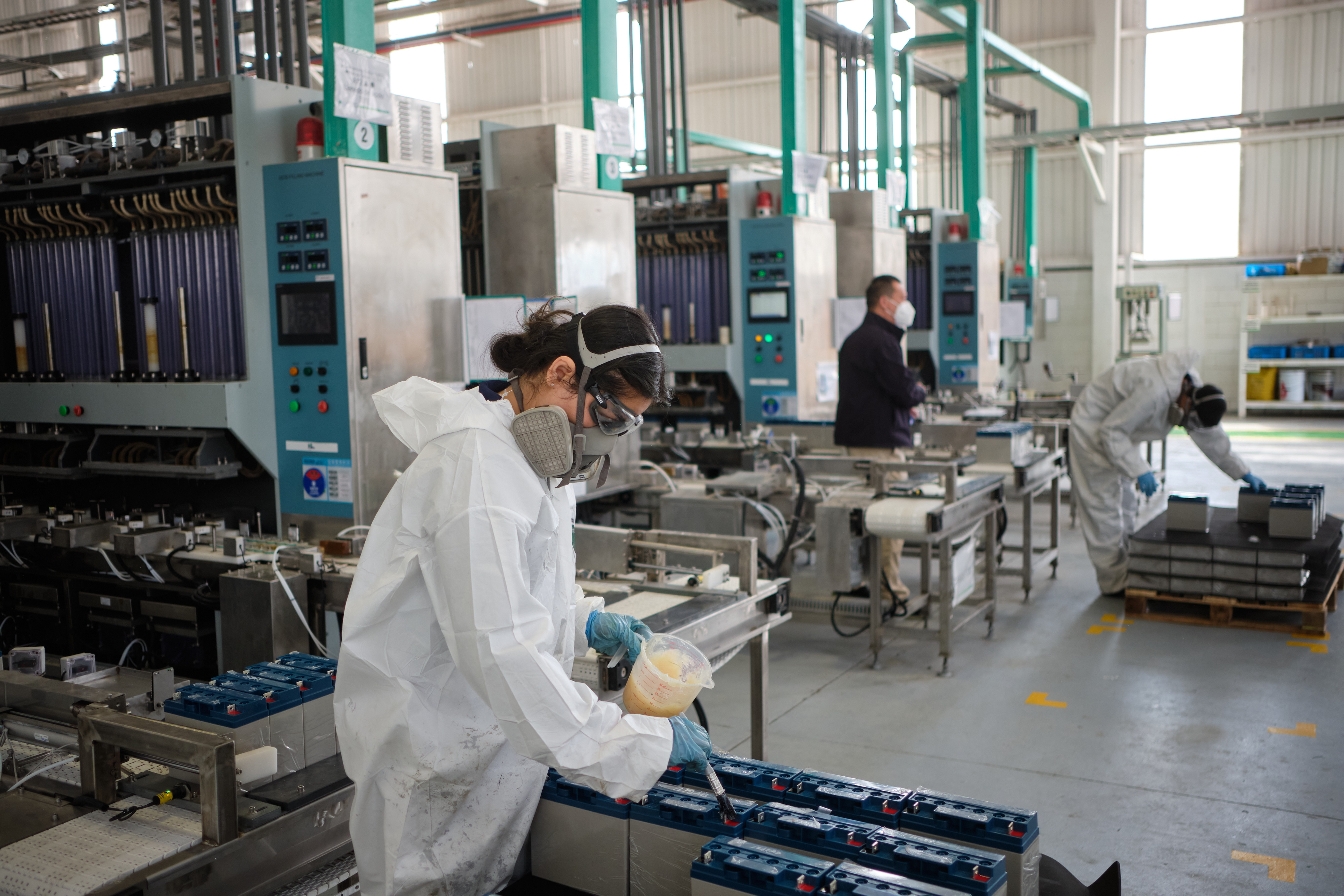 A worker wearing protective clothing and a mask applies vaseline to acid batteries at a factory in Coahuila, Mexico, with other employees in the background