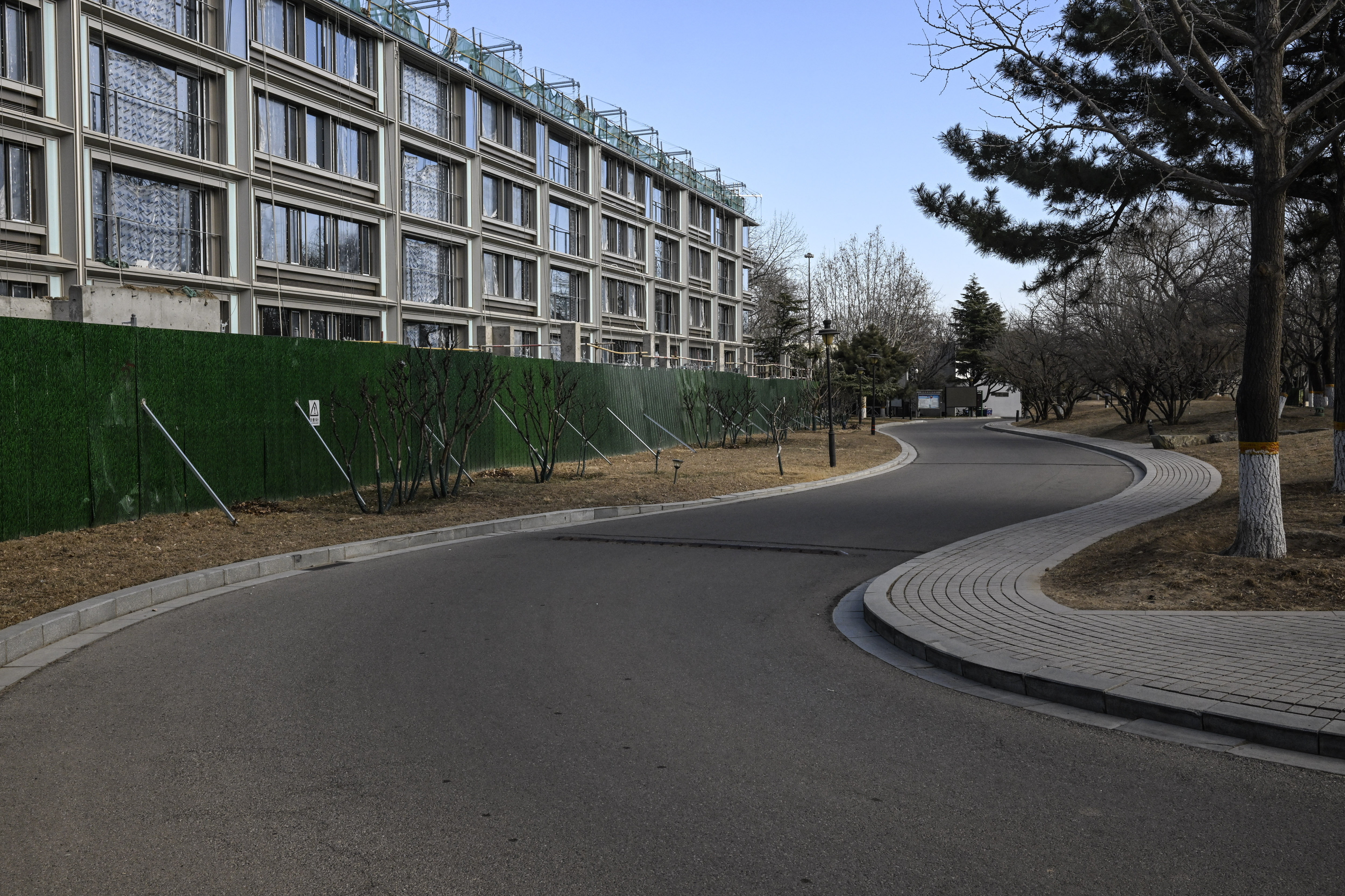 A winding road curves around a modern residential complex with large windows and balconies. A green construction fence runs along the left side, partially obscuring the lower part of the buildings.