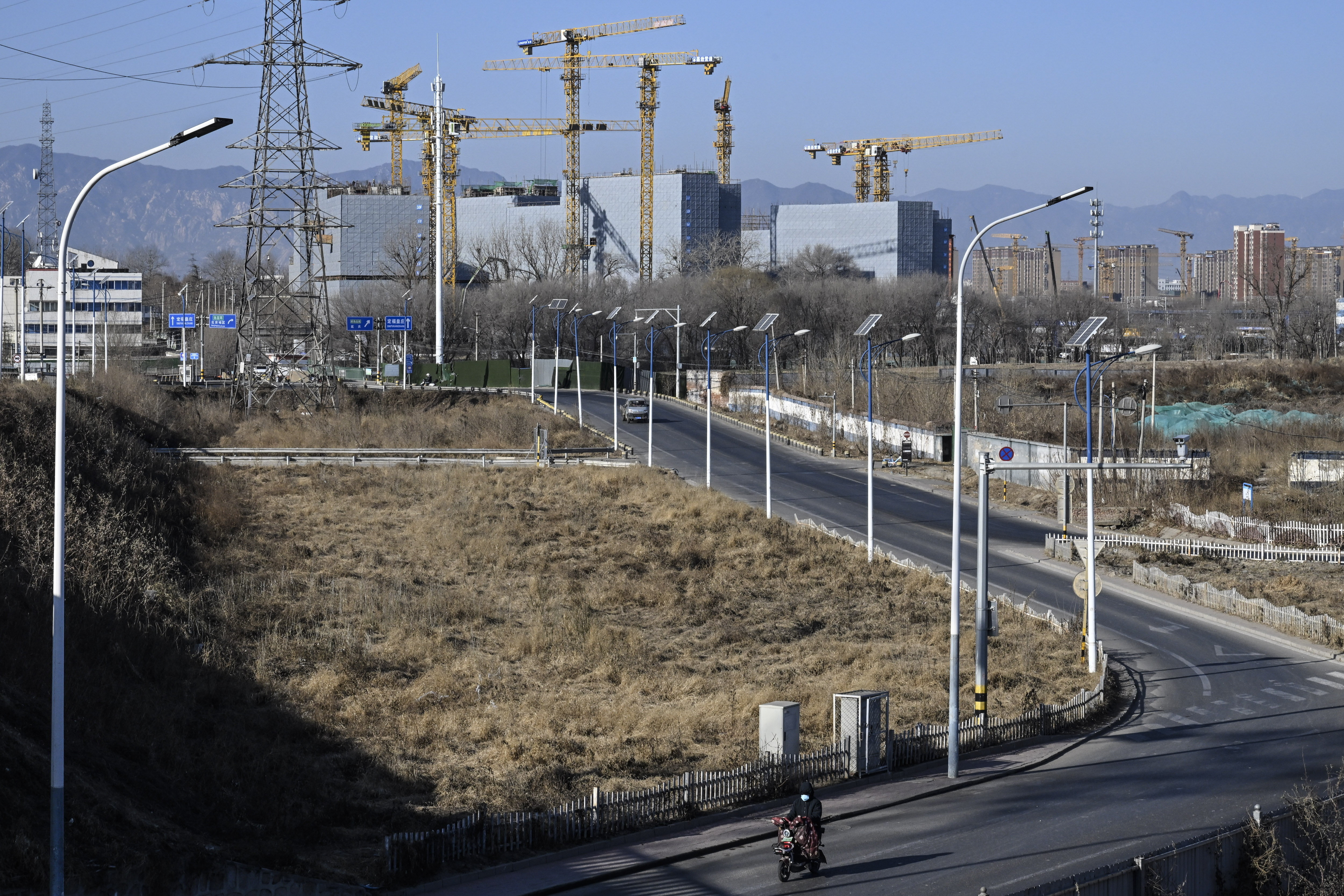 A wide road with streetlights runs alongside a barren patch of land, with a motorbike rider traveling along the road. In the background, multiple construction cranes loom over partially built high-rise buildings, with mountains visible in the distance.