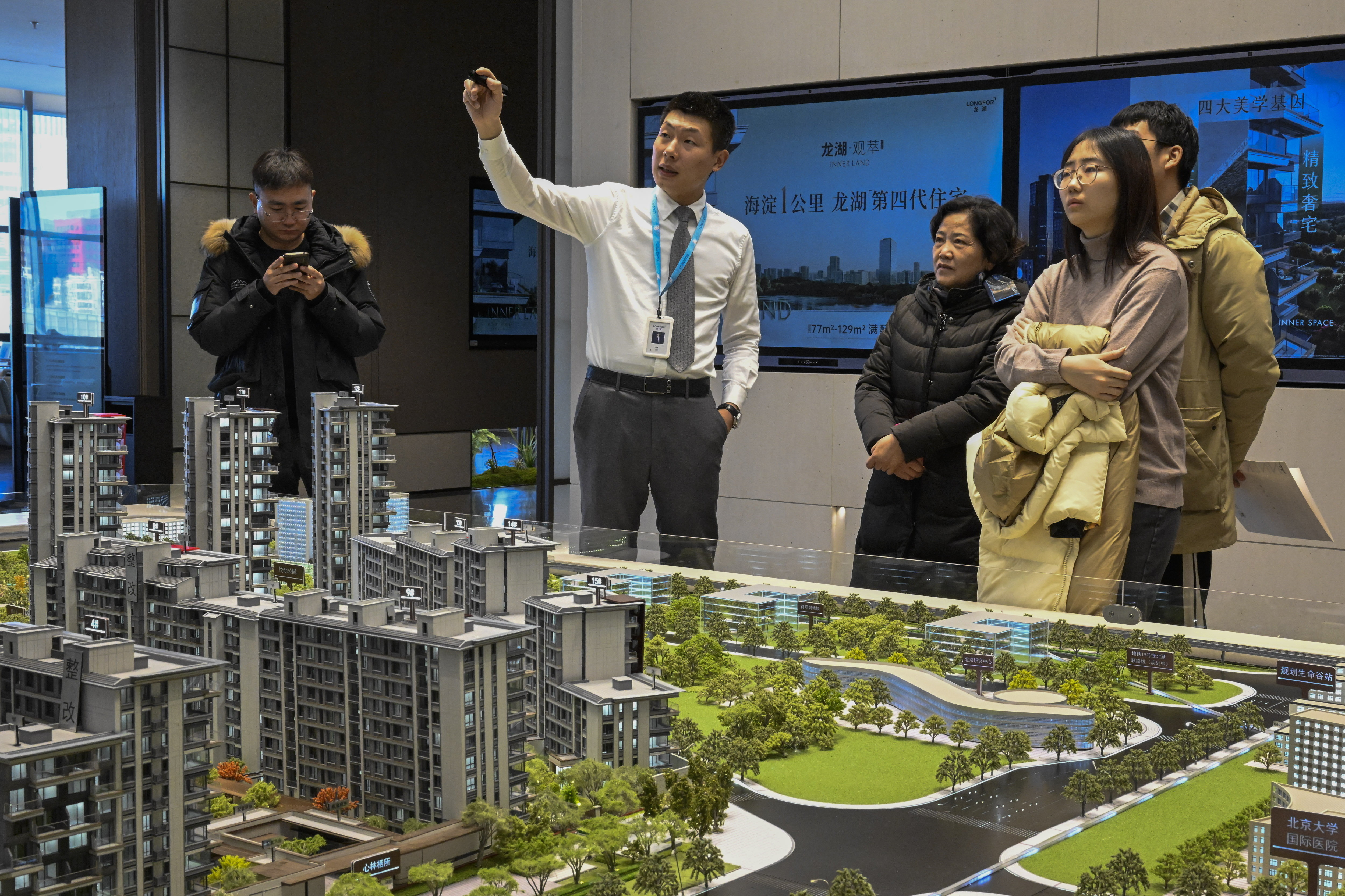 A man in a white shirt gestures while explaining a scale model of a real estate development to a small group of people. The model features clusters of high-rise buildings, green spaces, and roads. Large screens in the background display real estate information.