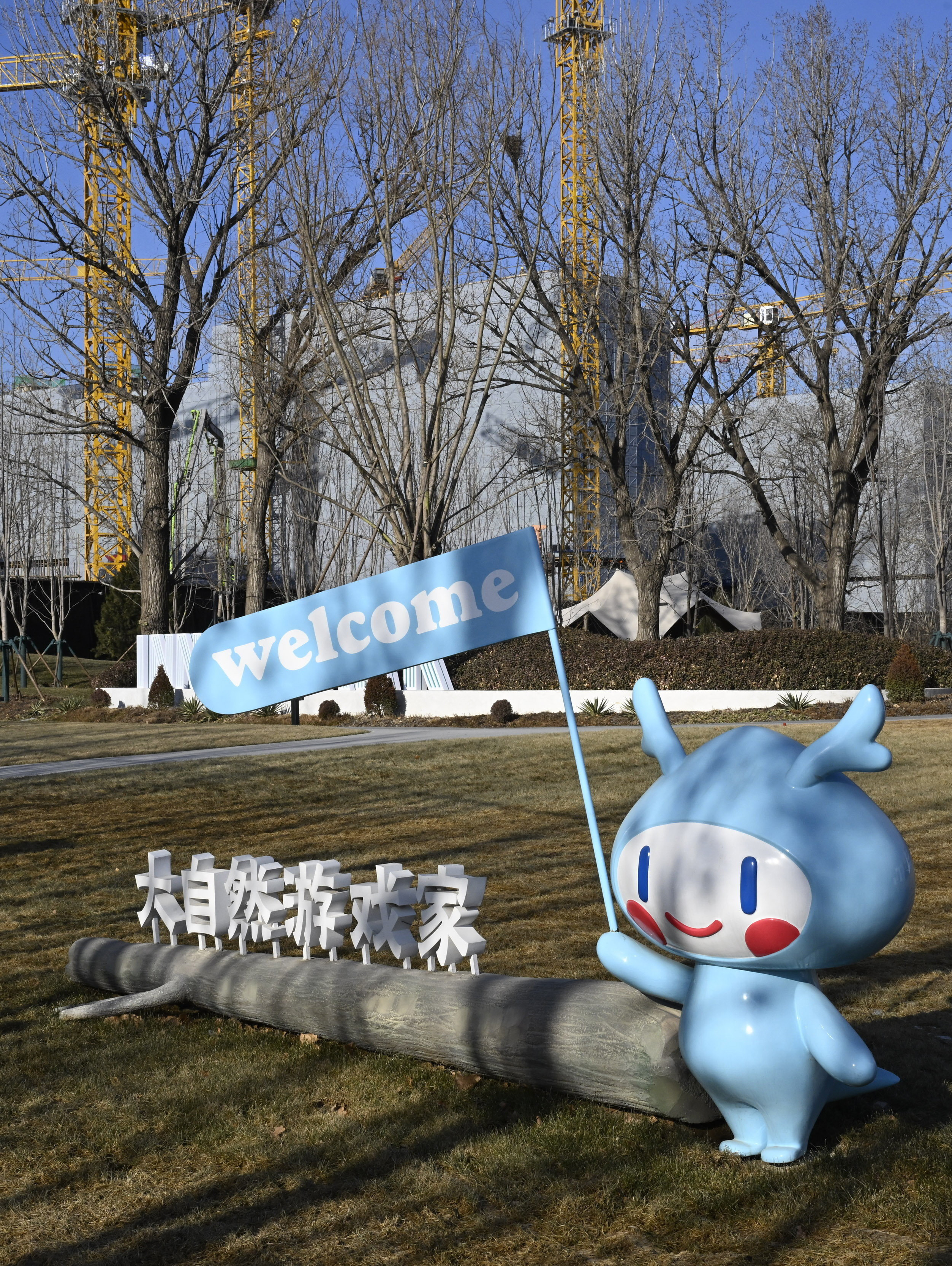 A blue cartoon-like statue holds a flag that says "welcome". The statue is on a grassy area with a wooden log featuring Chinese characters. In the distance, several construction cranes and shells of large apartment buildings are visible.