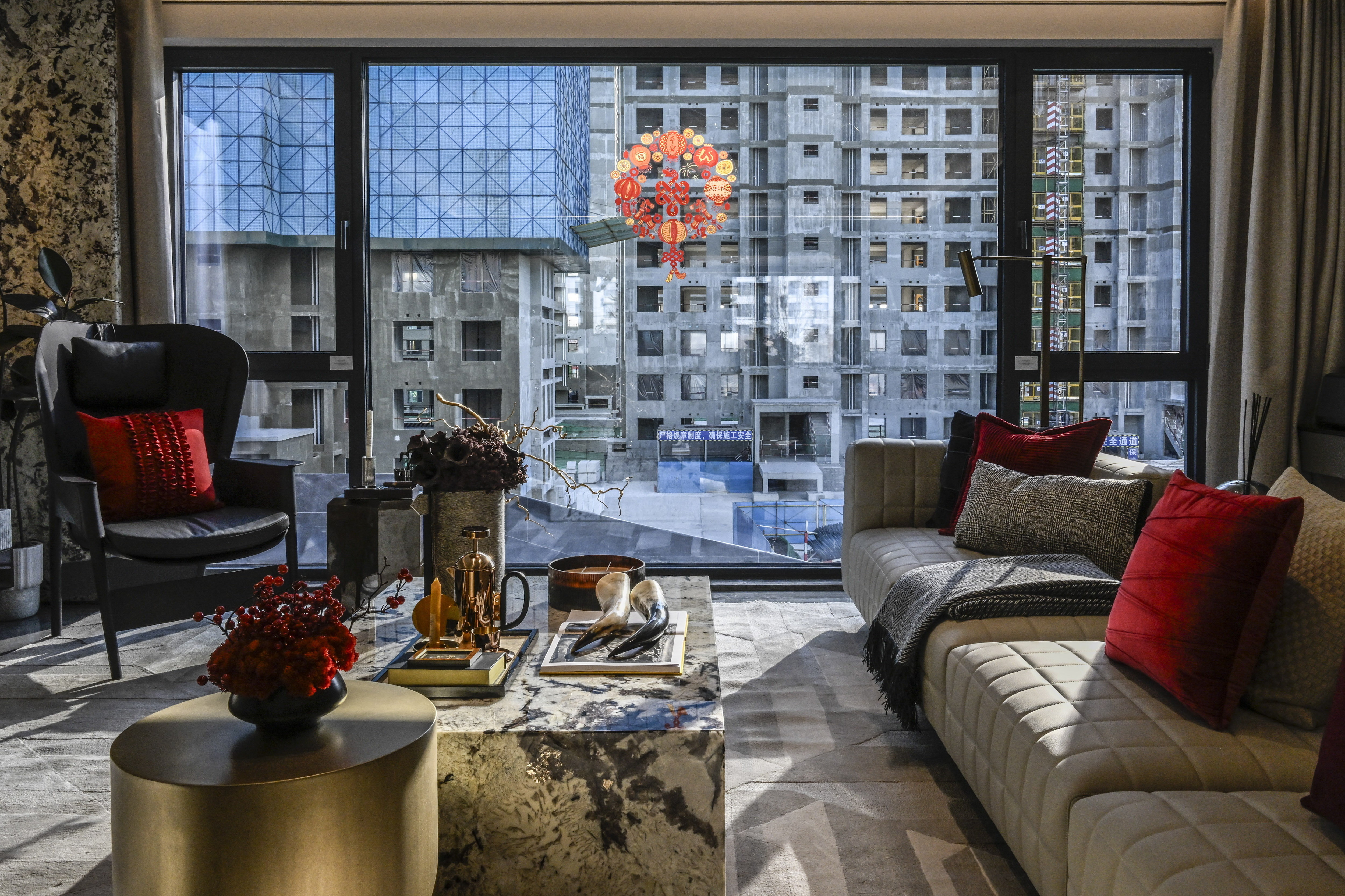 A modern, well-furnished living room with a large floor-to-ceiling window overlooking a construction site with unfinished high-rise buildings. The room features a beige sofa with red accent pillows, a marble coffee table with decorative items, and a black armchair with a red cushion. A red Chinese New Year decoration hangs in the window.