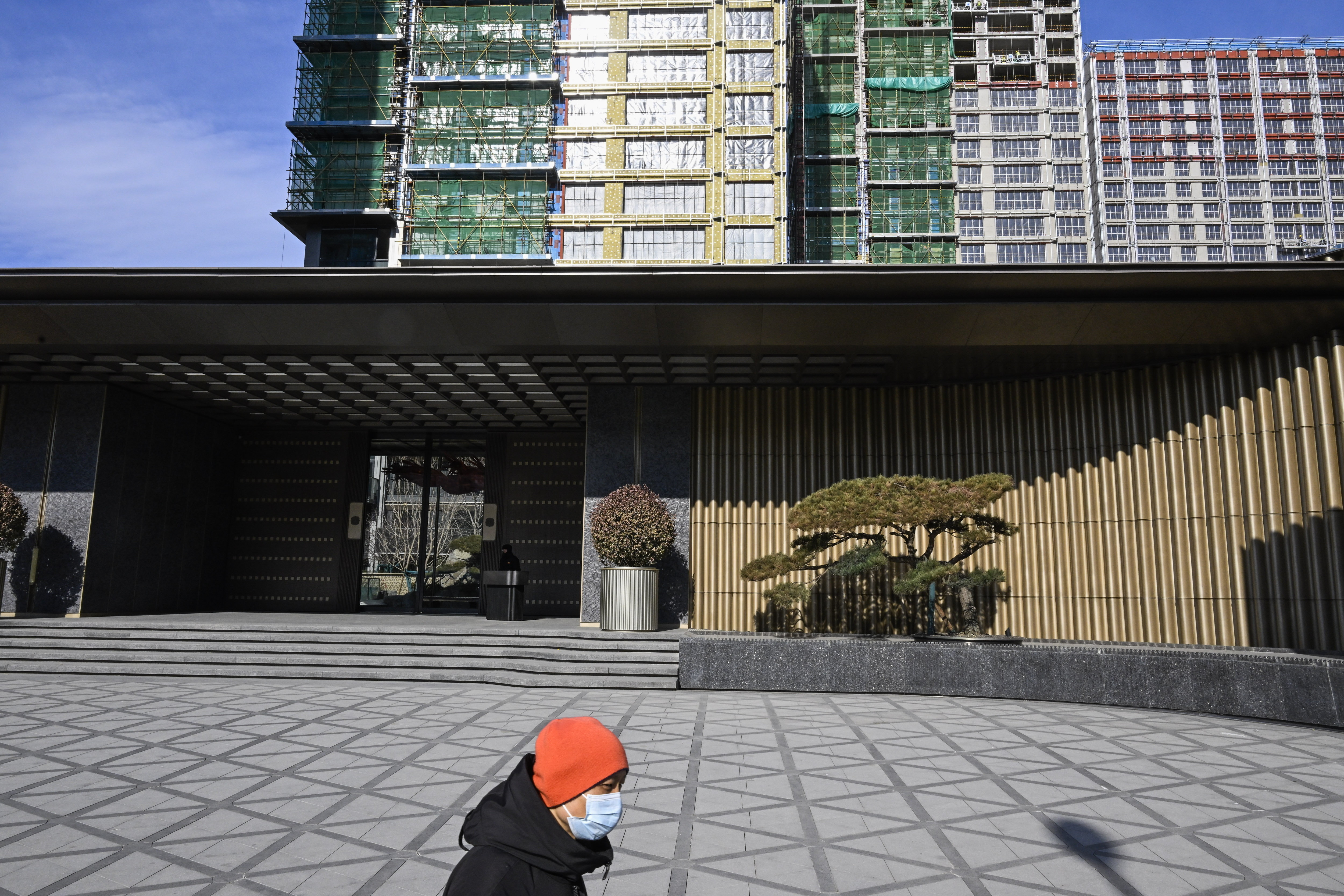 A large construction site with multiple high-rise buildings in various stages of completion. Several cranes tower above the site, and scaffolding covers parts of the buildings. In the foreground, two people with a yellow bicycle cross a wide, empty road.