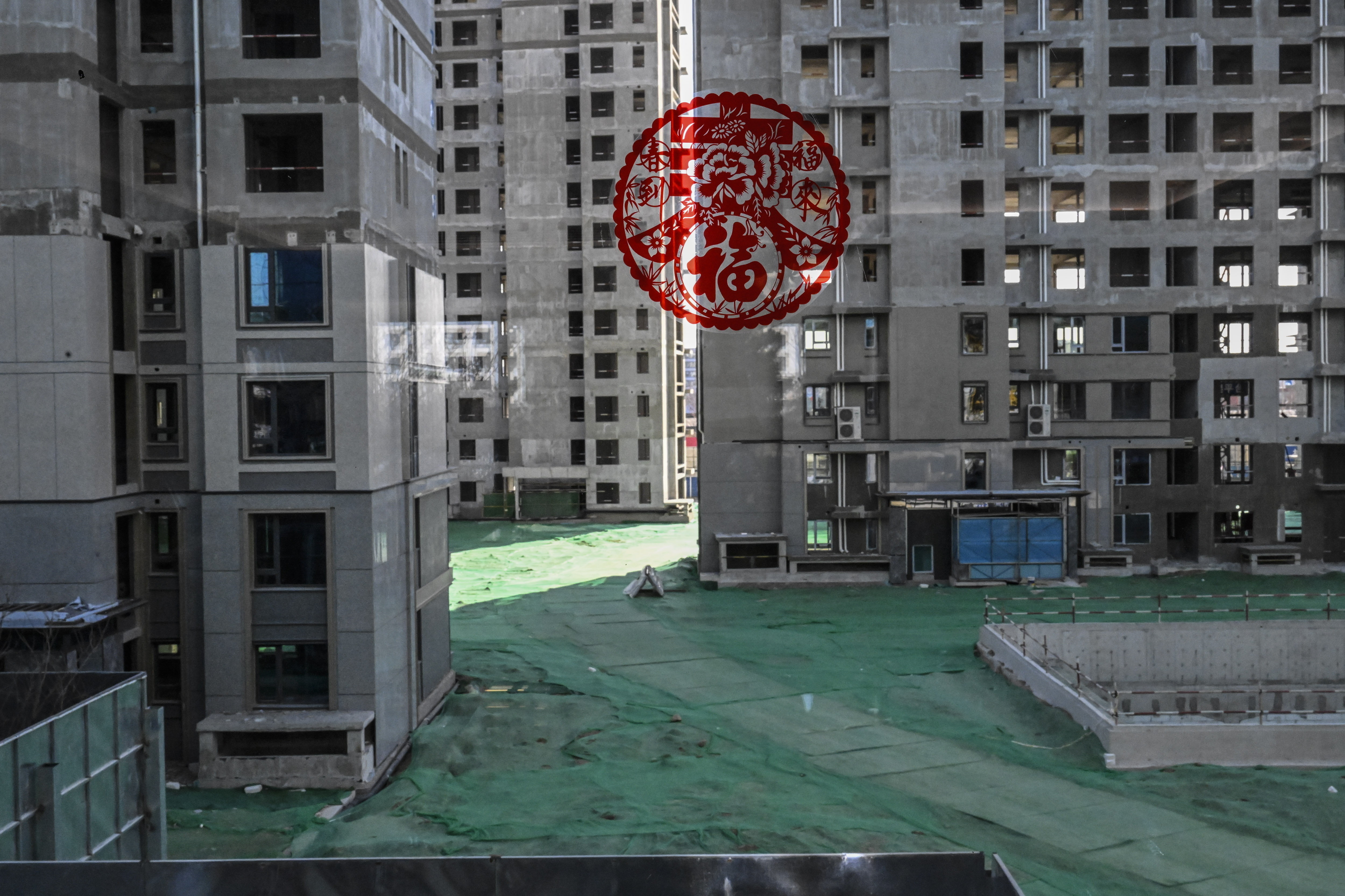 A view from a window overlooking a construction site with multiple unfinished high-rise apartment buildings and the ground is covered with green protective netting. A red Chinese New Year decoration is stuck to the window.