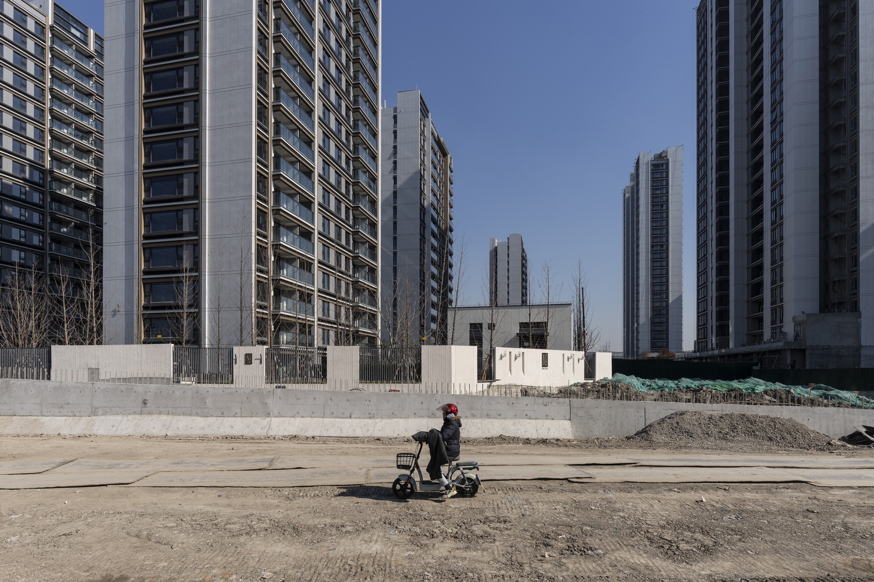 A person wearing a red helmet rides a scooter across an unfinished road in front of a construction site. In the background, tall modern residential buildings with glass facades contrast against a blue sky.