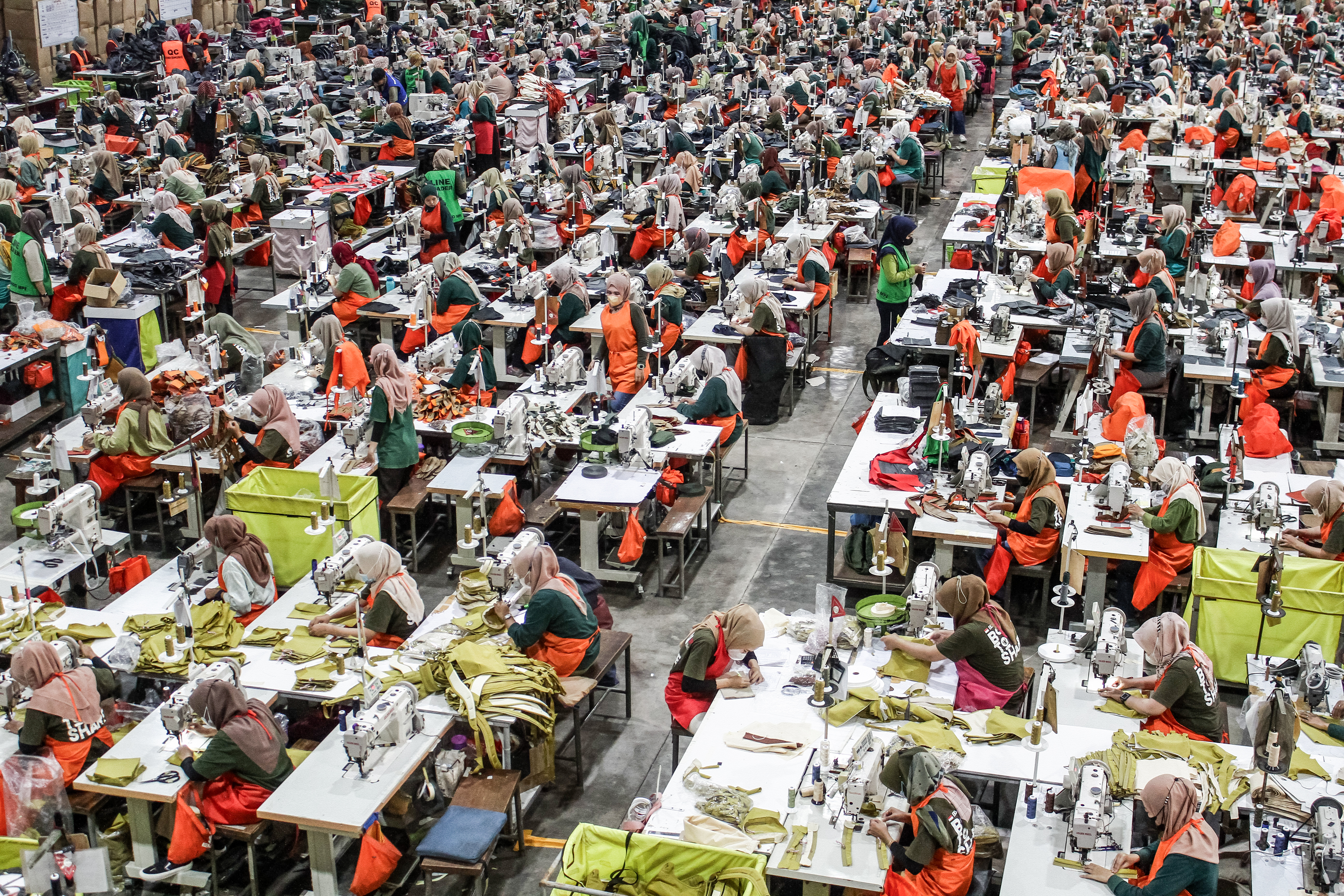 A large garment factory in West Java, Indonesia, filled with rows of workers, mostly women wearing headscarves and orange aprons, seated at sewing machines. The factory floor is densely packed with workstations, fabric pieces, and sewing materials, creating a vibrant and busy atmosphere of mass textile production.