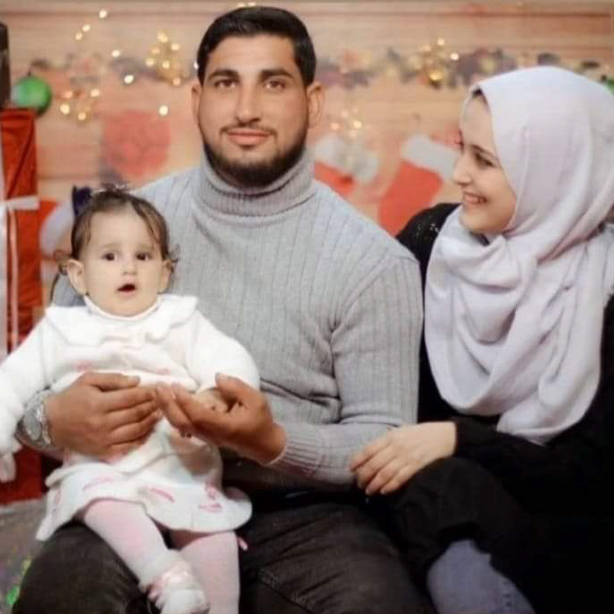 A photo of a smiling family, with a man holding a baby girl on his lap while a woman looks adoringly at her.