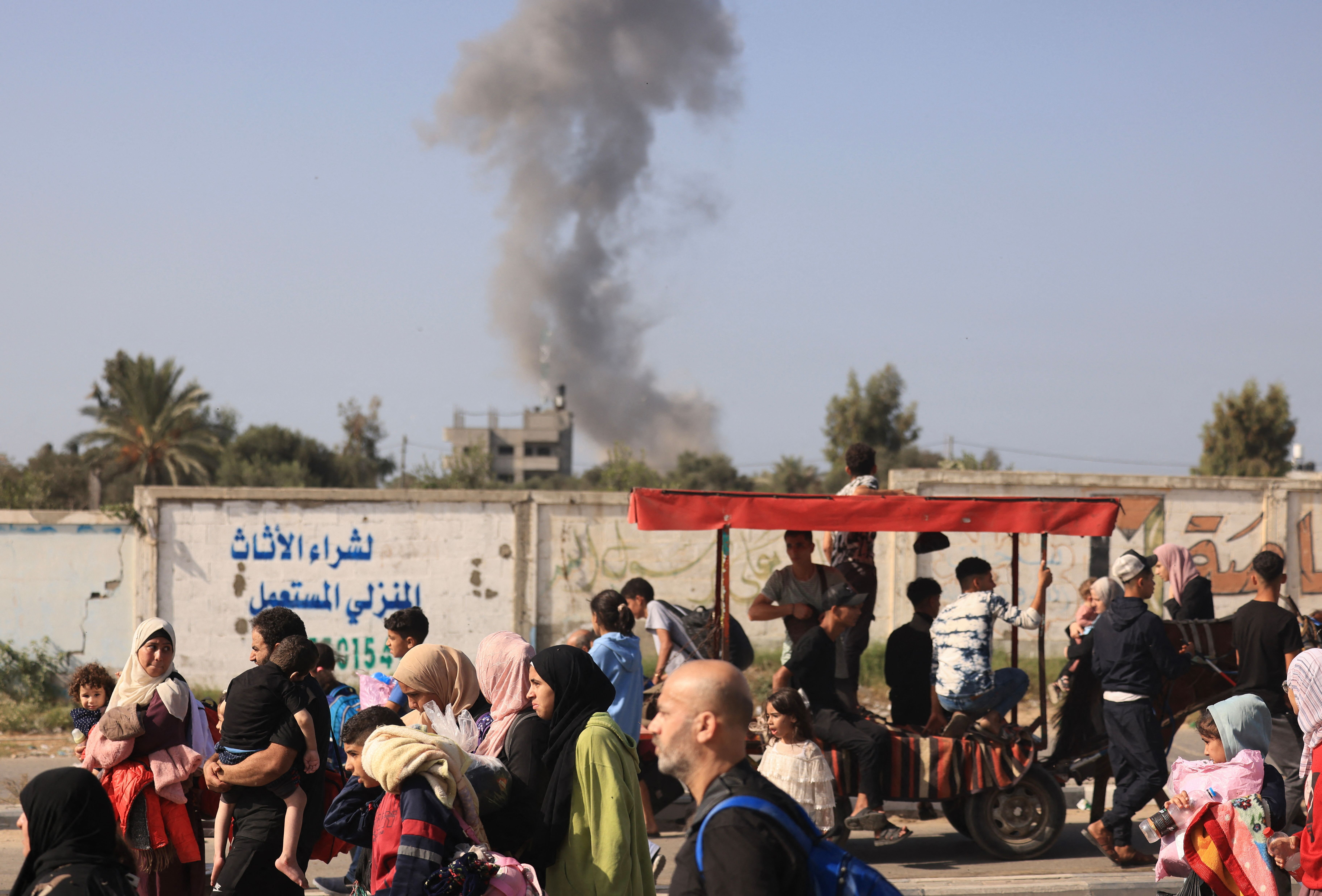 Families, some with children in their arms, walk along a street next to a wall covered in graffiti. Smoke can be seen in the distance.