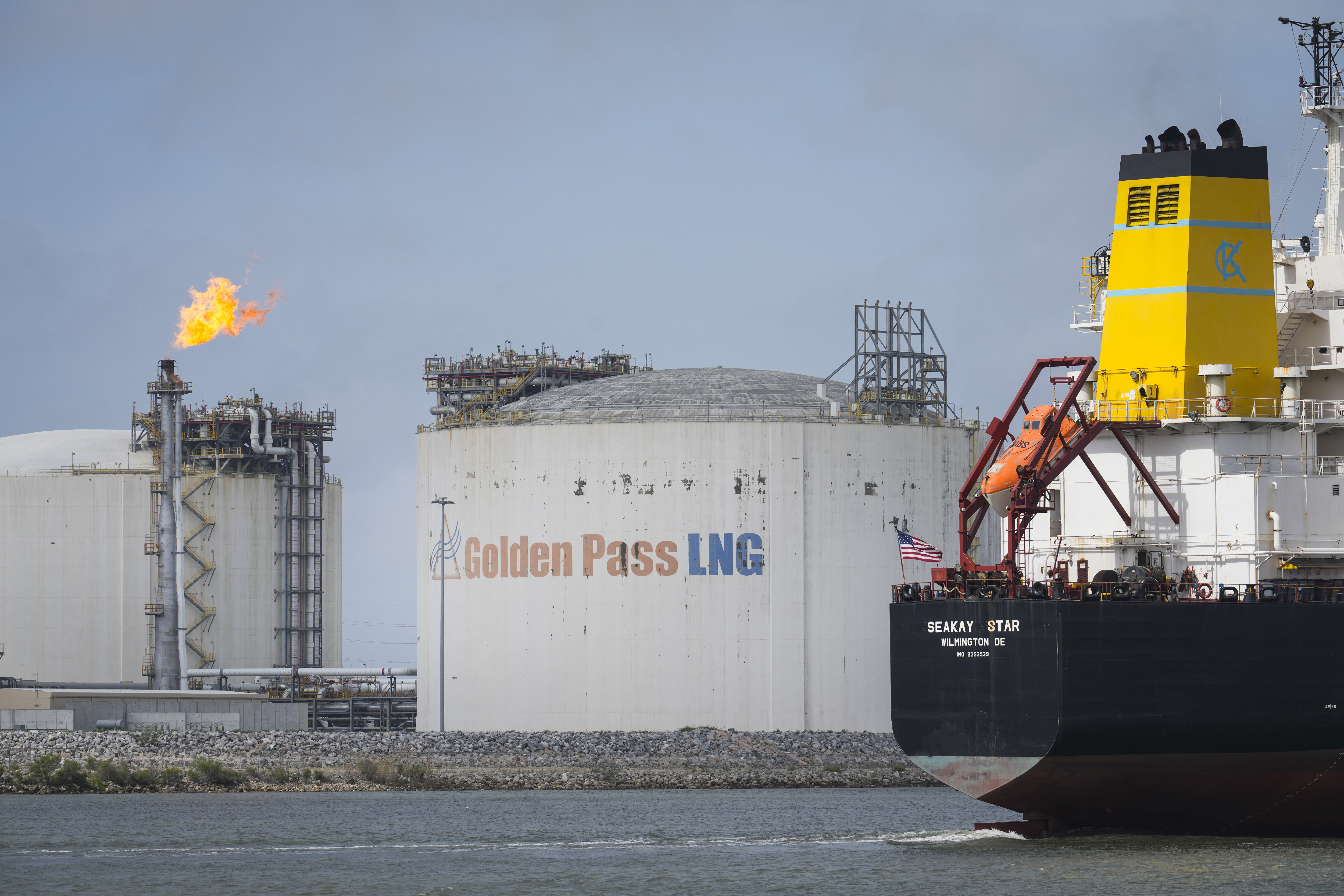 A large tanker passes an LNG export terminal with white storage tanks labelled “Golden Pass LNG” and a flare burning in the background at Port Arthur, Texas.