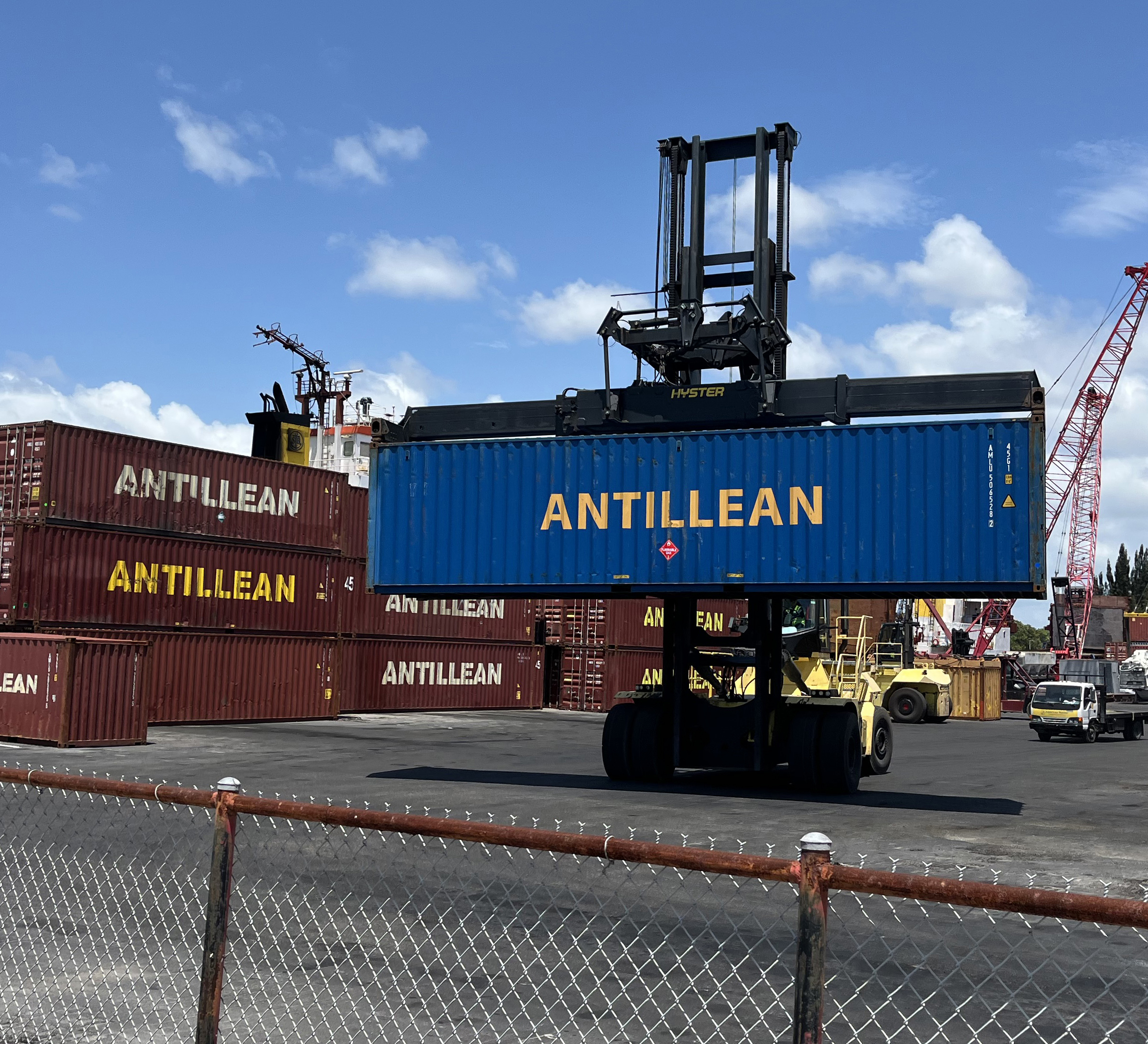 A large blue shipping container labeled "Antillean" is being moved by a yellow container handler in a freight yard. Several red containers, also marked with "Antillean," are stacked in the background under a clear blue sky. A chainlink fence is in the foreground