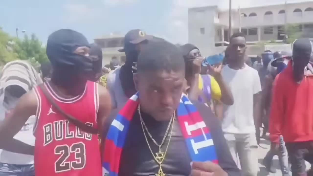 A group of people, some wearing masks, walk outdoors in a daytime street setting, with one individual in the foreground draped in a red, blue, and white scarf.