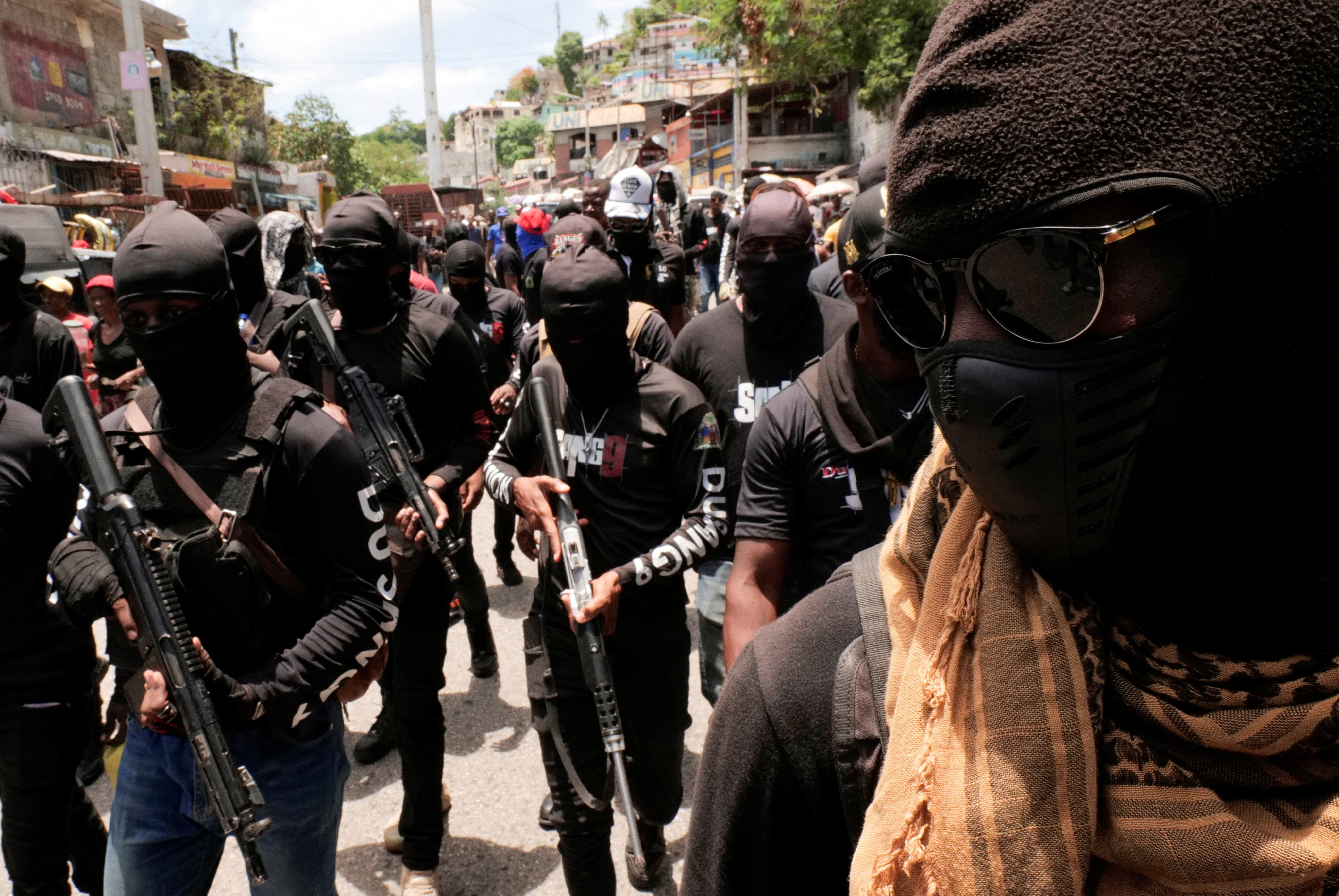 A densely packed group of masked individuals dressed in matching black clothing and carrying rifles are marching down a street lined with buildings and trees.