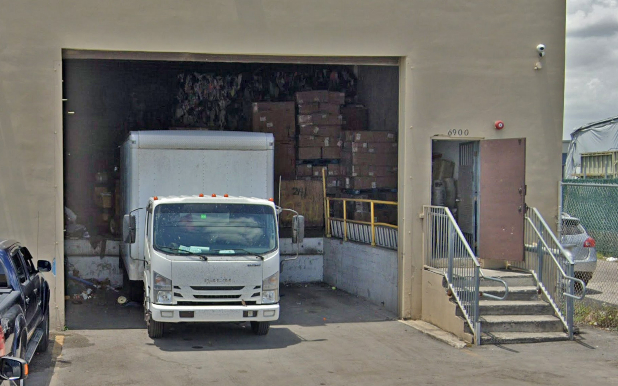 A white delivery truck is parked in front of a loading dock filled with stacked cardboard boxes, alongside a compact staircase and an open door leading into a light yellow coloured warehouse.