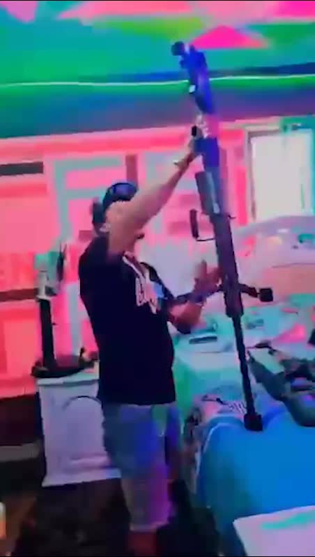 A person in casual clothing stands in a room with colorful, neon lighting, holding a large rifle. The background shows a bed and a window.