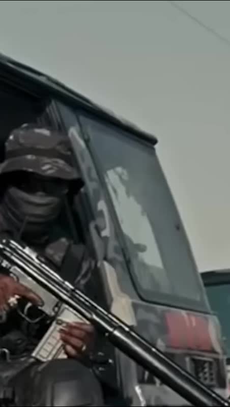 A camouflaged man wearing tactical gear and a face covering is holding a rifle while seated in a military-style vehicle.