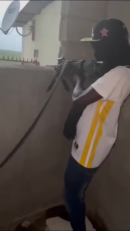 A person stands in a corner wearing a cap and mask, pointing a belt-fed machine gub out of a window.