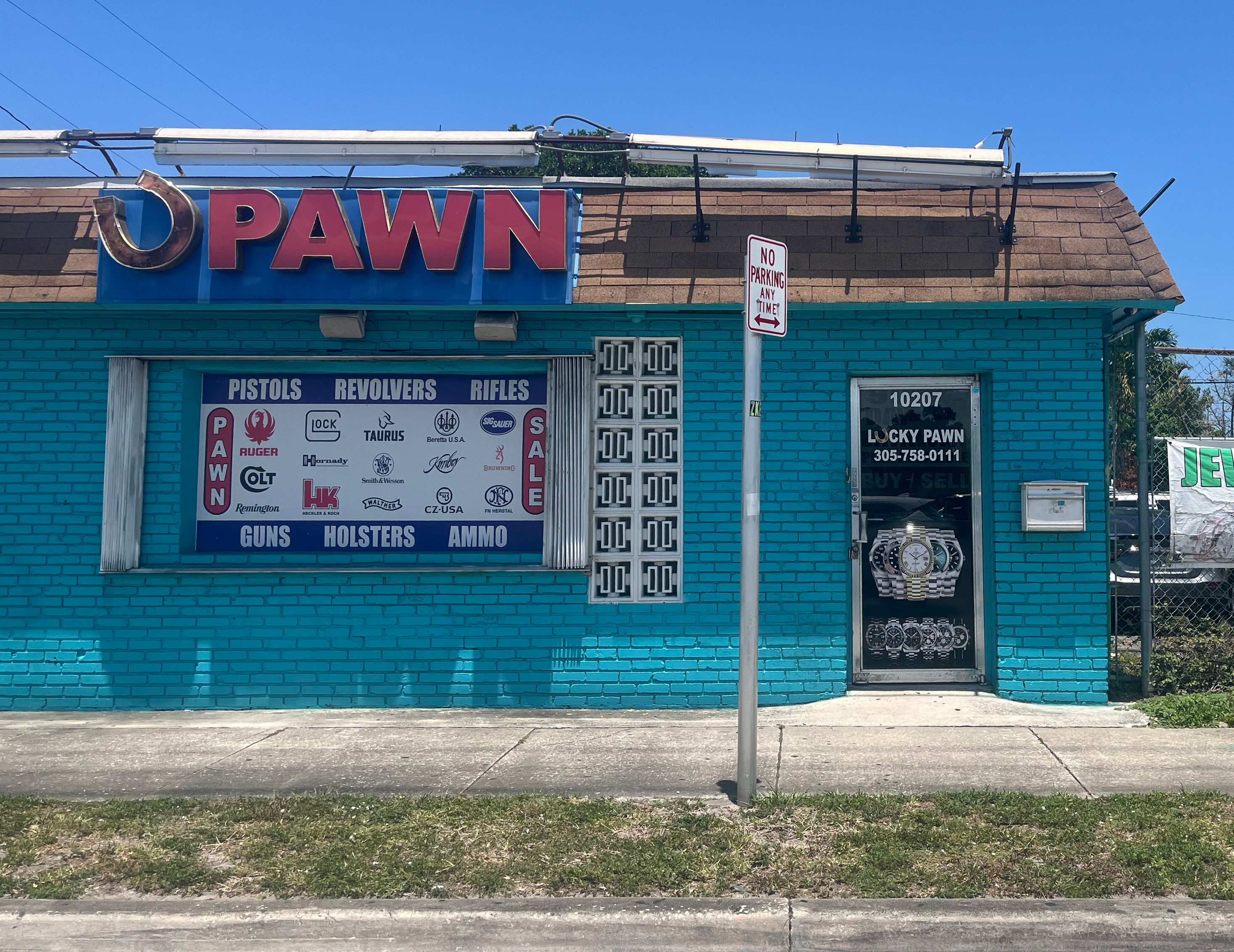 A turquoise low-rise brick building with a large sign with a horseshoe and sign that reads "PAWN" in red letters above an advertisement for pistols, revolvers, and rifles. The entrance door displays the name "Lucky Pawn" along with a phone number, and a "No Parking" sign is visible in front.