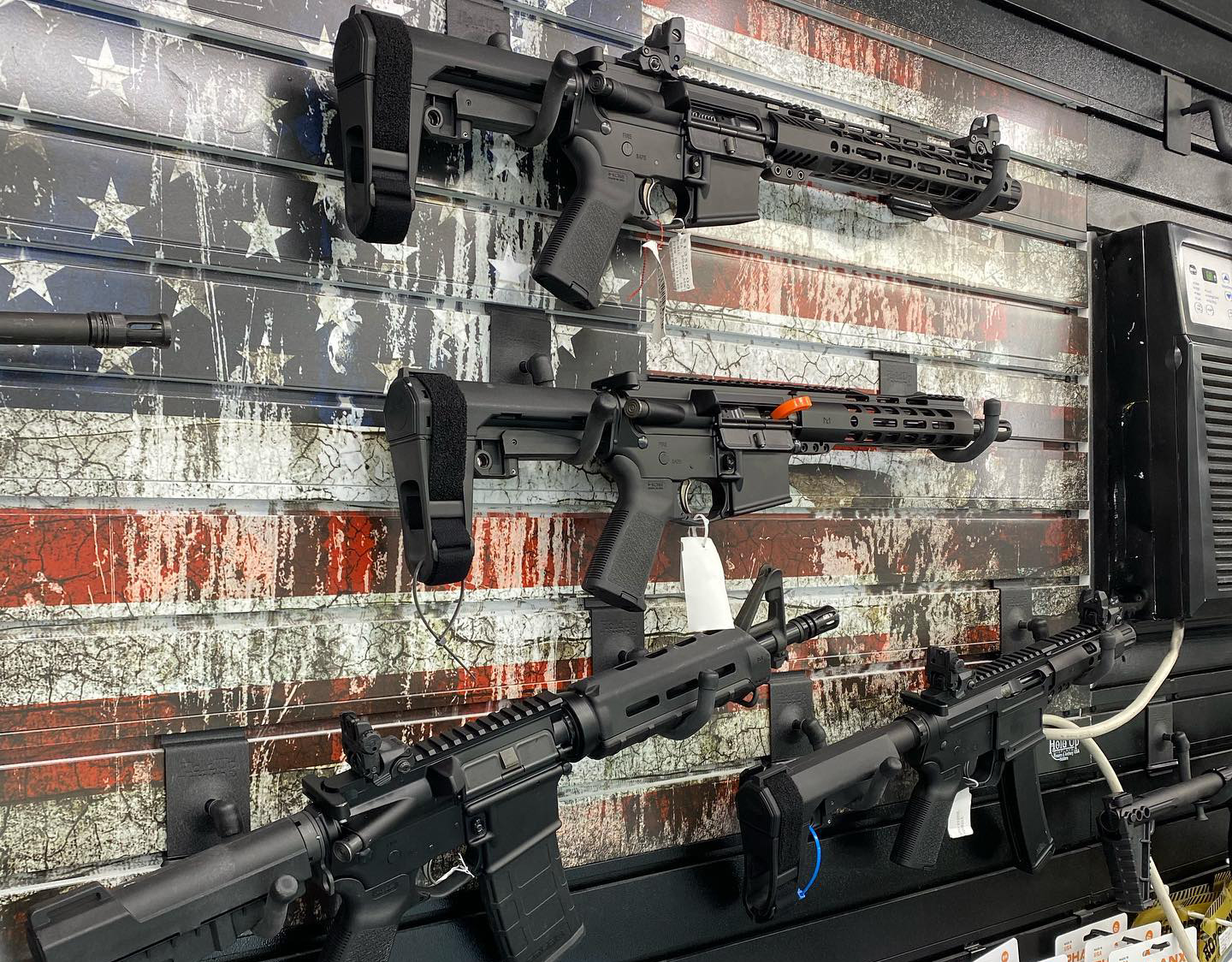 A display wall showcases several semi-automatic rifles mounted on racks, against a weathered American flag backdrop. The firearms have sales tags attached.