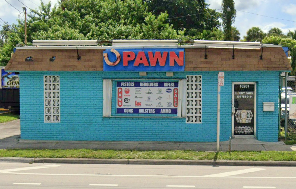 A small, turquoise brick pawn shop with a large horseshoe and "PAWN" sign above, advertising guns, holsters, and ammunition. The building features a variety of firearm brand logos and a side door with the shop's contact information.