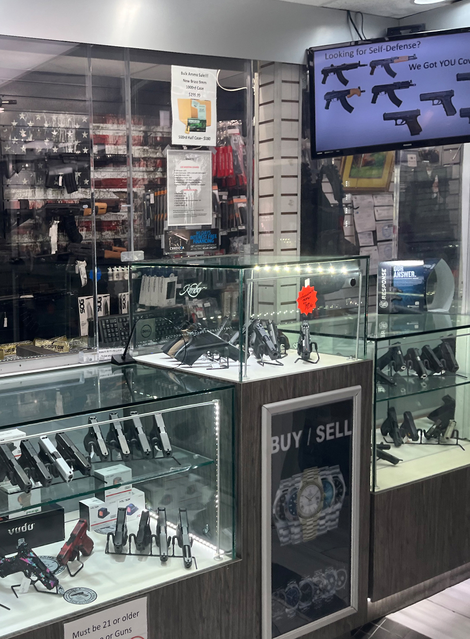 The image shows the interior of a pawn shop displaying various firearms in glass cases, with a sign indicating goods for self-defense and a digital screen showing different weapon models. There are promotional signs, including one for ammo and a warning that buyers must be 21 or older.