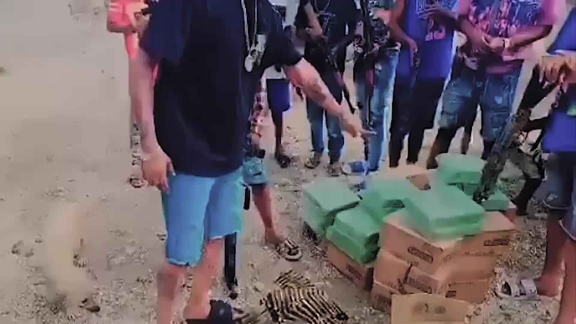 Group of people standing on a dirt surface, one man points to a collection of AK-style rifles leaning against a wall.