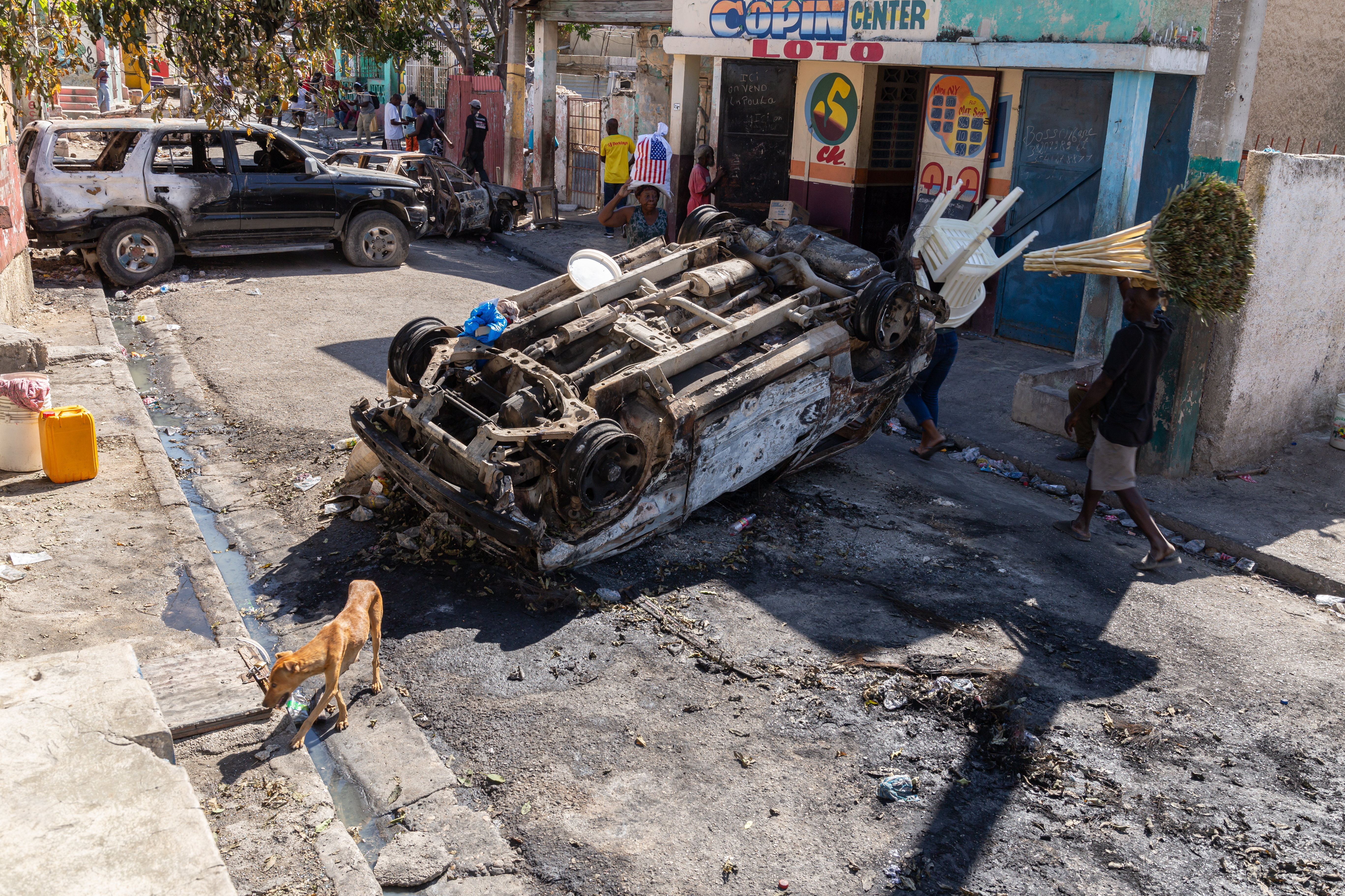 An overturned, burned-out car lies on a street with debris scattered around, while another charred vehicle is parked nearby. People walk past, one carrying chairs and another with a bundle on their head, as a dog sniffs around the gutter.