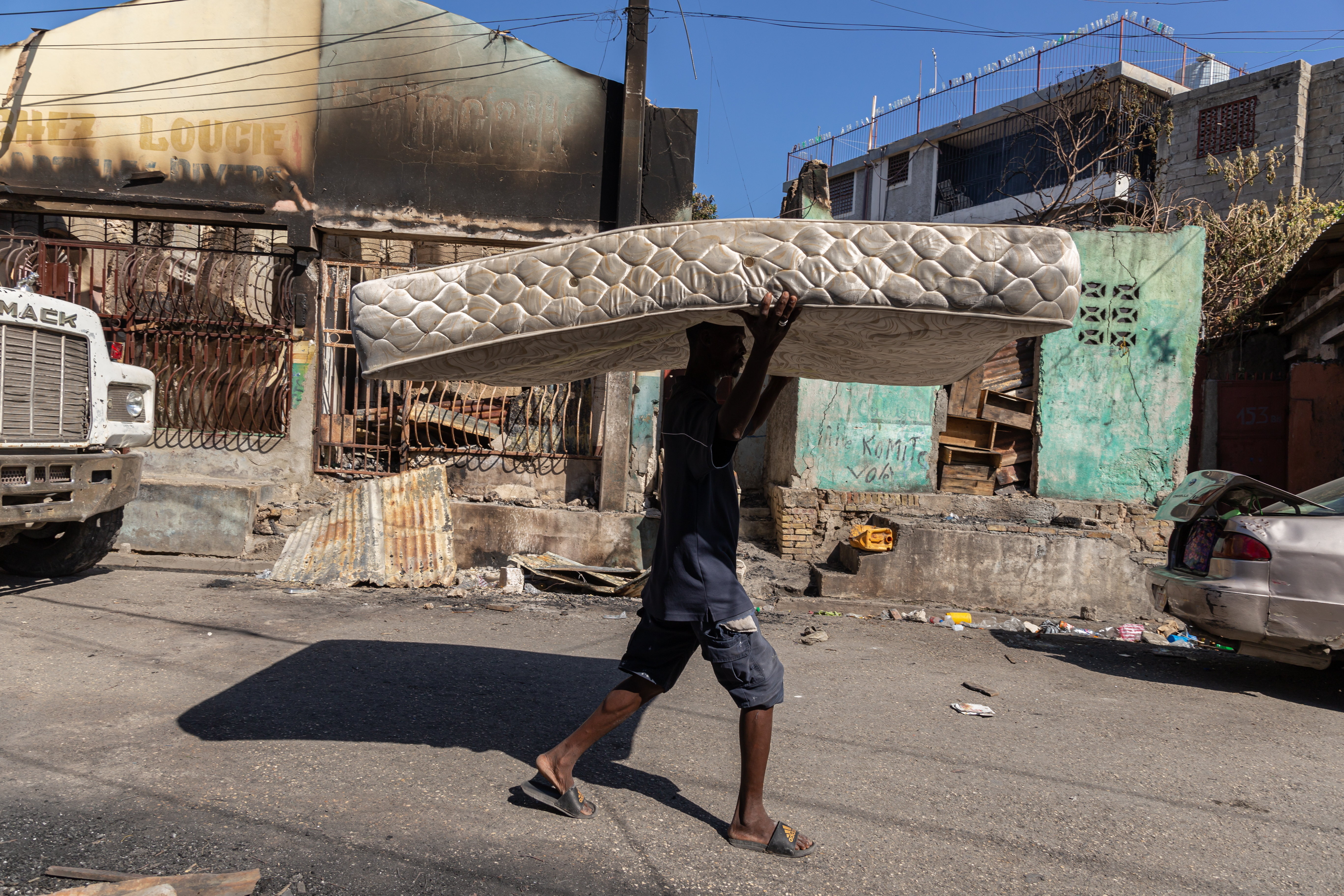 A person walks down a street carrying a mattress on their head, passing by a burnt building with peeling paint and litter scattered around.