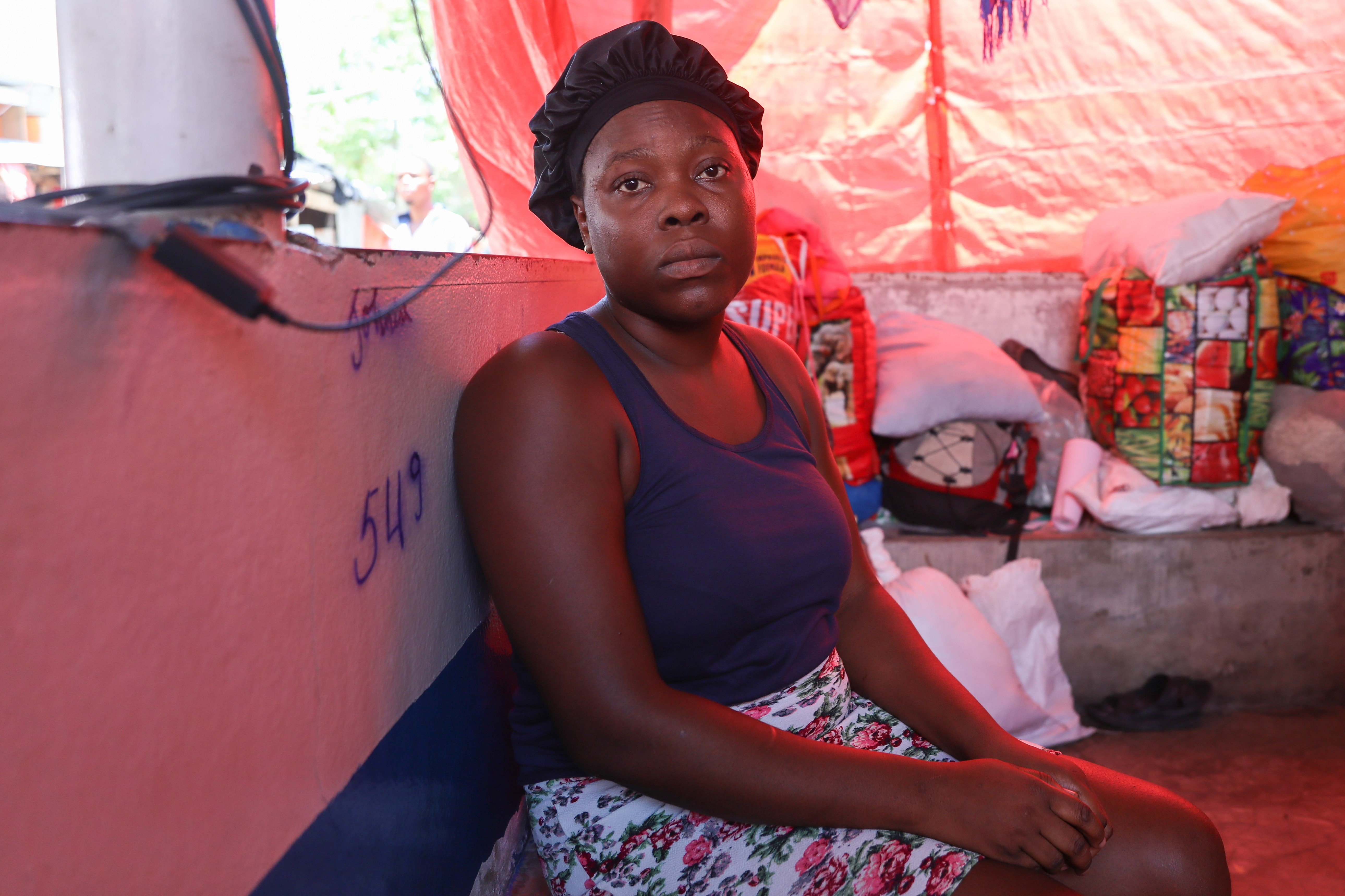 A woman sits against a pink painted wall inside a makeshift shelter with a red tarp roof. She looks directly at the camera, surrounded by colorful, packed bags and supplies.