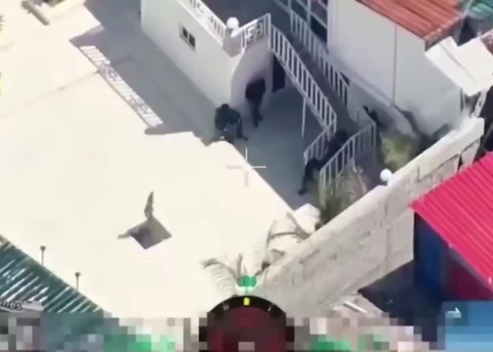 Aerial view showing several people near a building with white stairs and a red roof, captured through a camera crosshair overlay. The people run inside just before an explosion happens where they were standing.