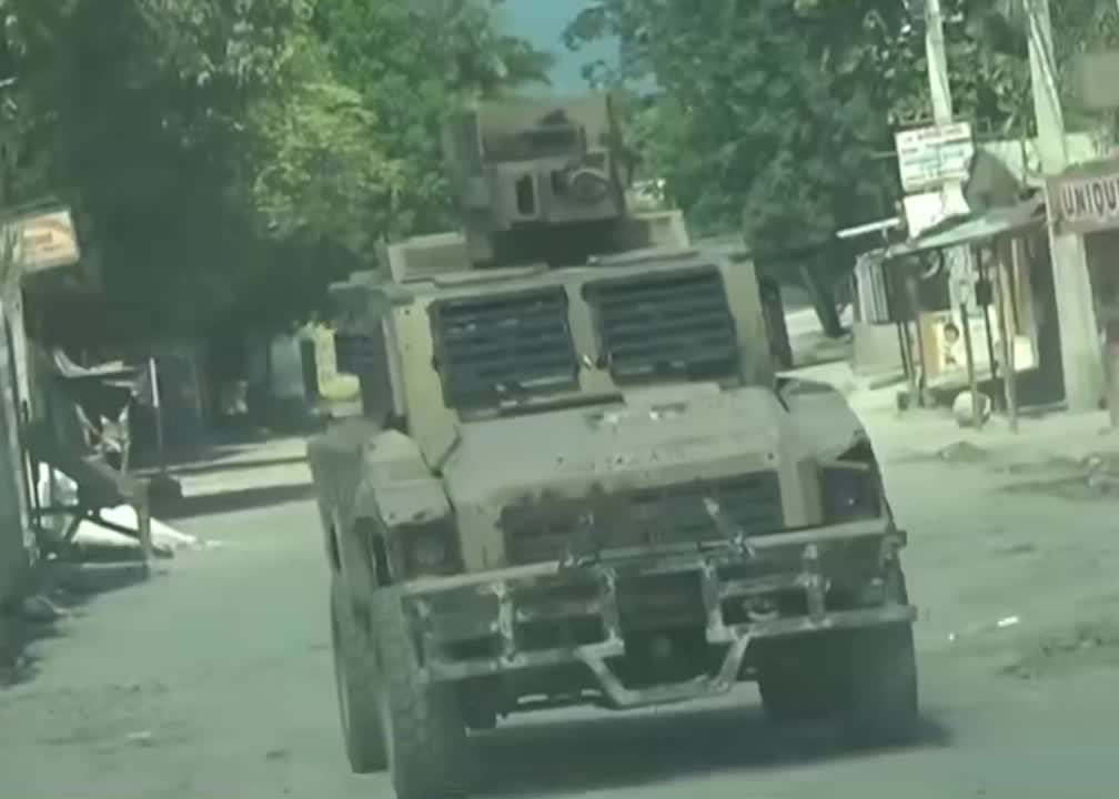 A armored vehicles is driving around deserted streets lined with small shops and trees. Policeman are inside the vehicles pointing rifles out.