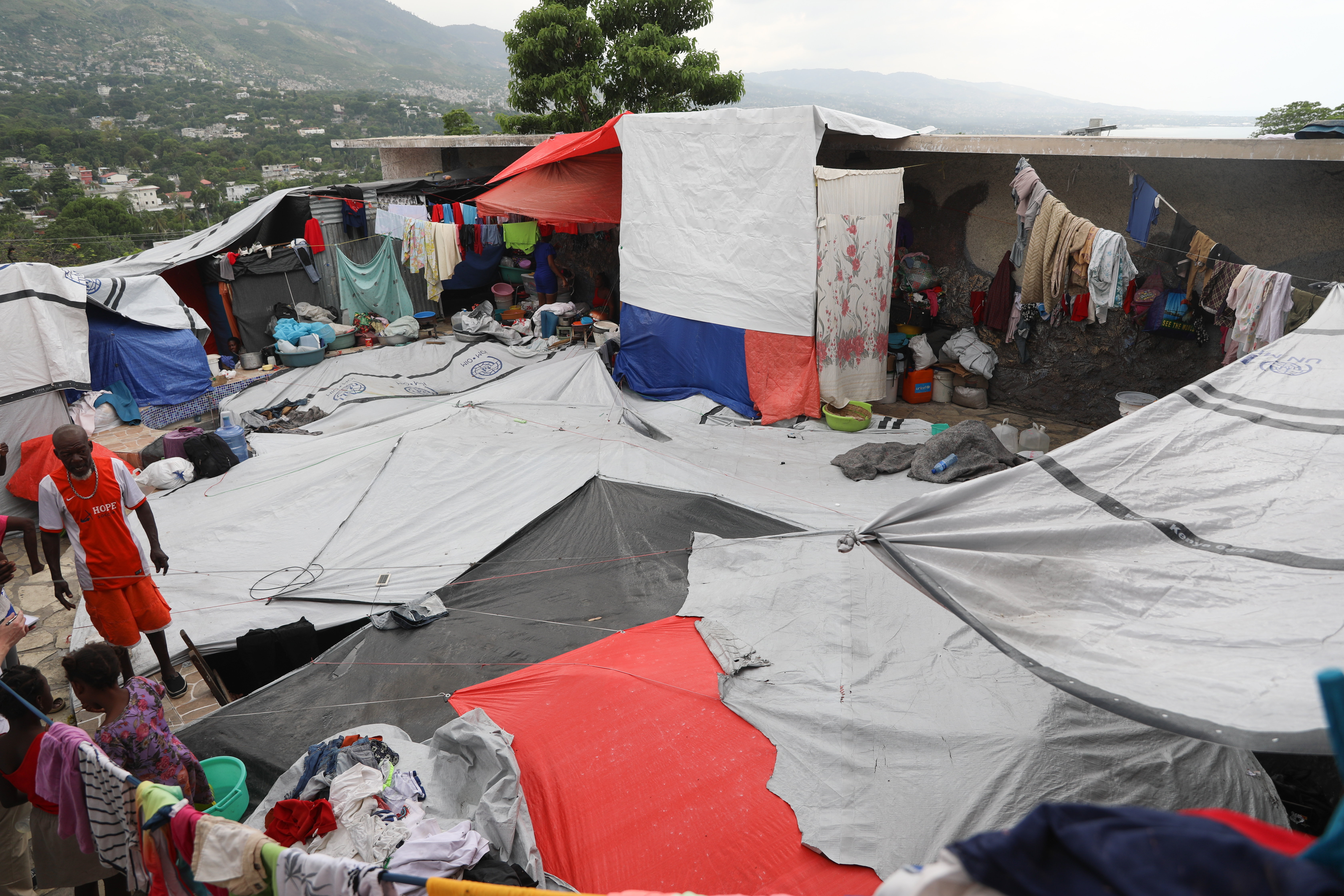 On the roof of a building is a collection of makeshift shelters created from tarps and cloths, with people seen moving around and clothing hanging to dry. Hills and buildings are in the distance.