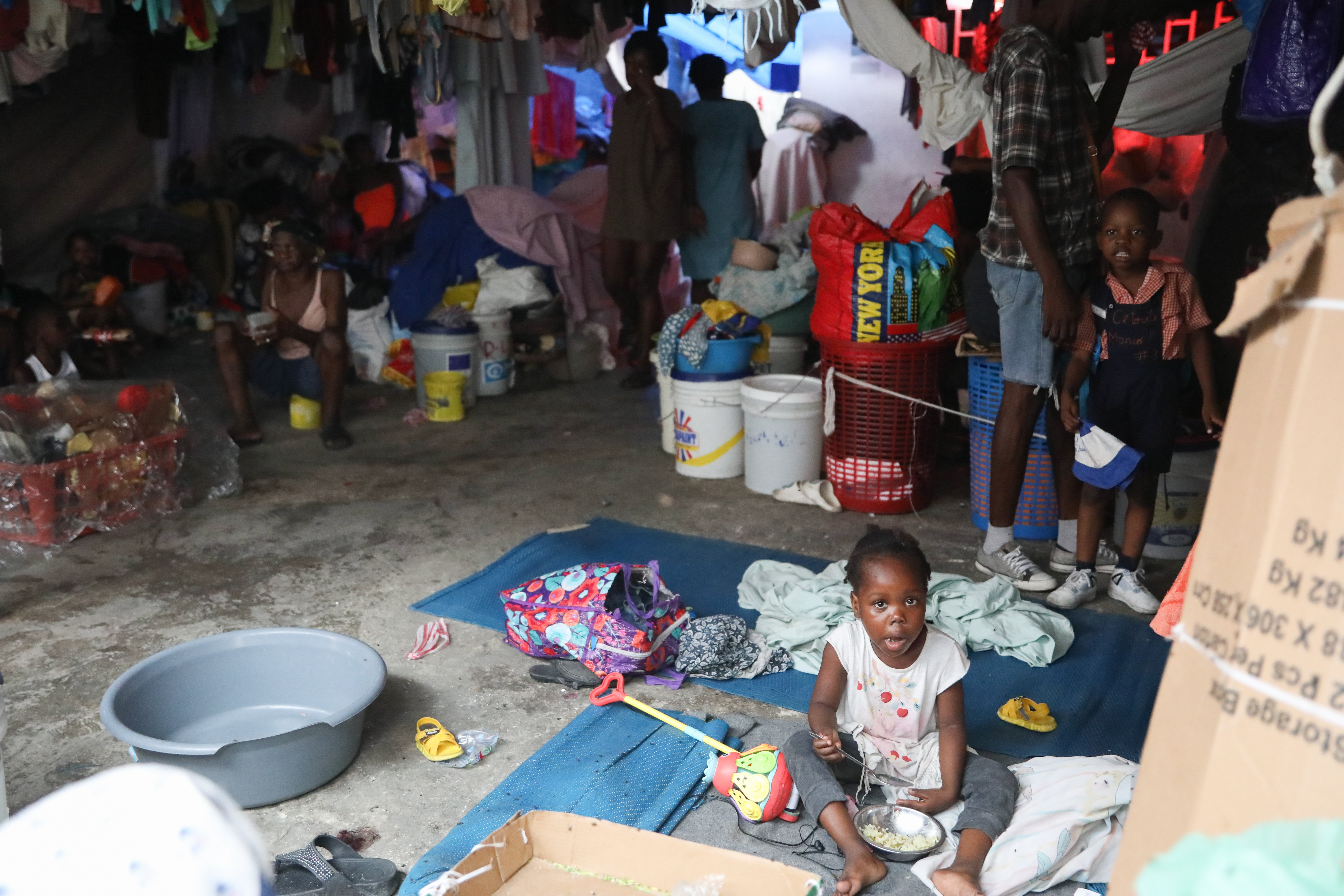 A dimly lit room crowded with people and belongings, where a young child sits on the floor eating from a bowl amidst scattered items like toys and clothes. In the background, various individuals are engaged in different activities, surrounded by bags, buckets, and hanging laundry.
