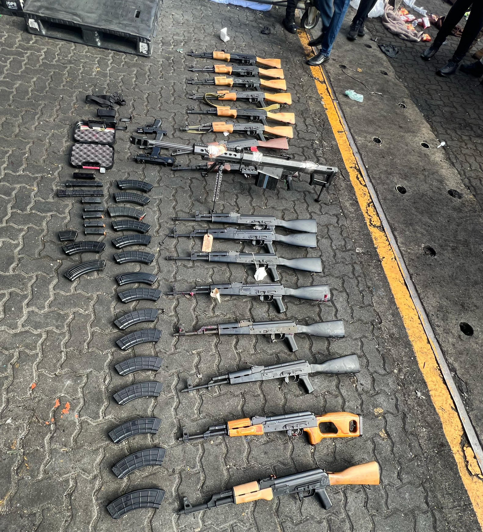 A collection of firearms and ammunition magazines are laid out on a paved ground, with rifles arranged in rows and multiple magazines aligned next to them.