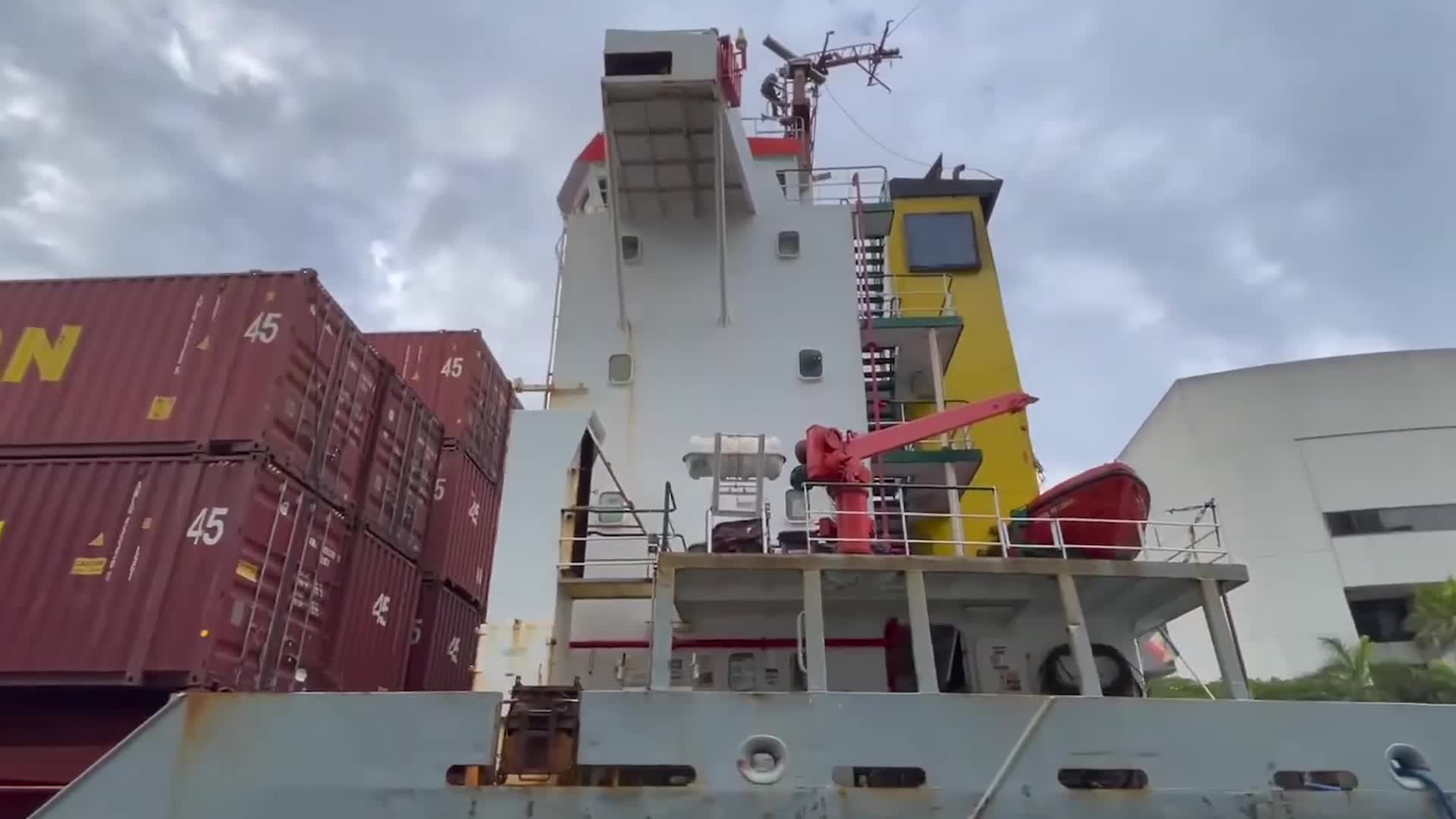 A large cargo ship with stacked shipping containers and the vessel's control tower, set against a cloudy sky.