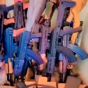 A collection of AK-style assult rifles are leaning against a wall. They are predominantly dark-colored with curved magazines.