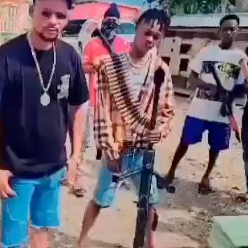 A group of men stand outdoors, with one individual prominent in the foreground holding a large rifle with a belt of bullets strung over his shoulder. The men are wearing shorts and t-shits and in the background is a vehicle and some buildings.