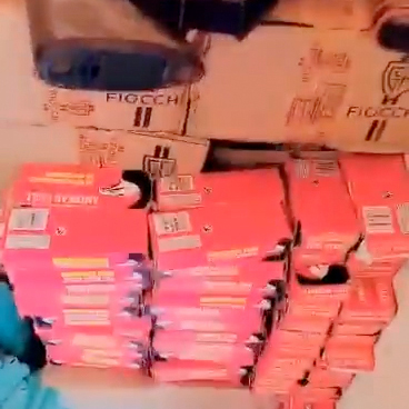 A large stack of red ammunition boxes is arranged against a wall, showing various labels and branding. The background consists of additional boxes, partially visible, with the brand name "Fiocchi" printed on them.