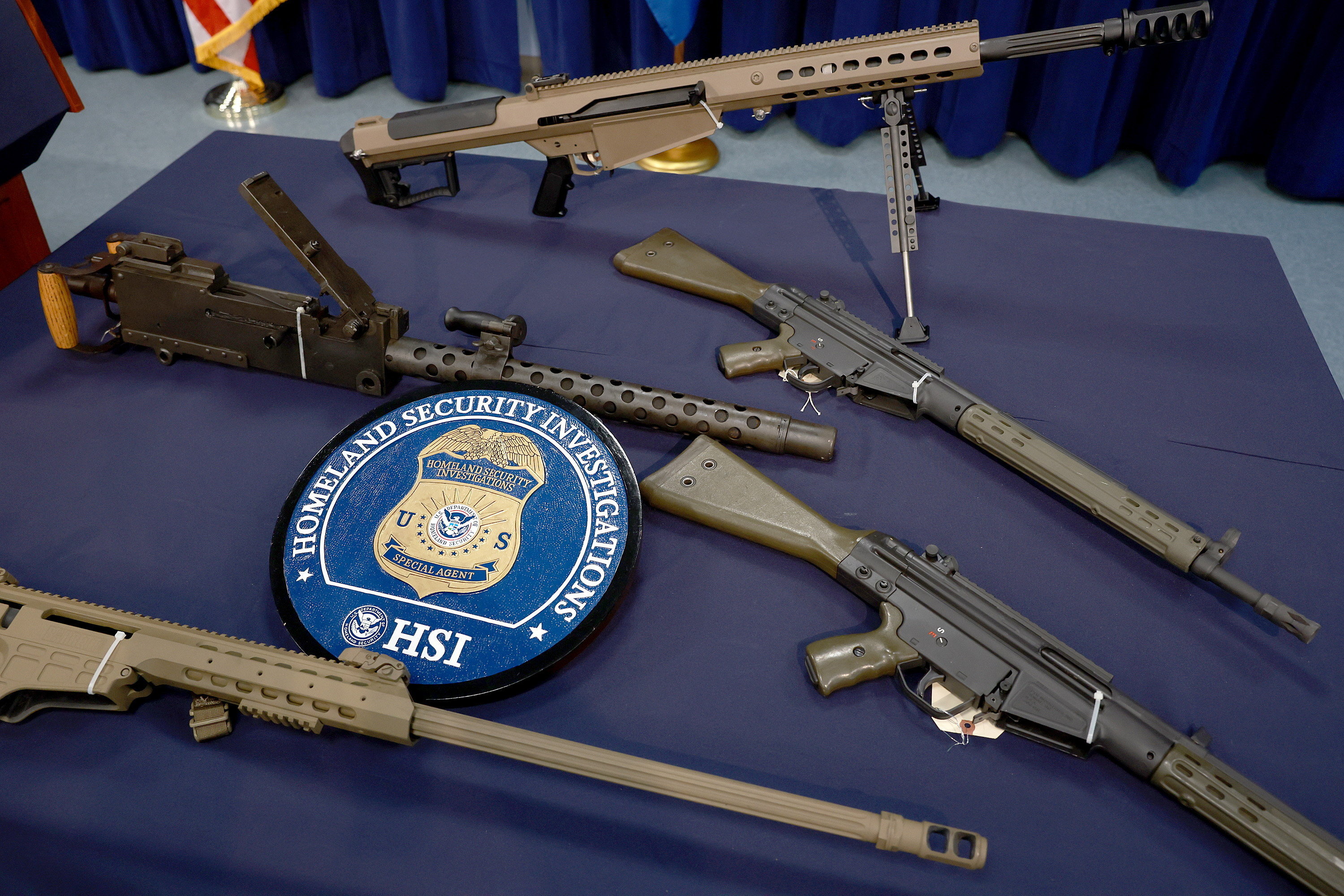 A collection of firearms displayed on a table, including rifles and machine guns, surrounds a Homeland Security Investigations emblem. The emblem is prominently placed among the weapons.
