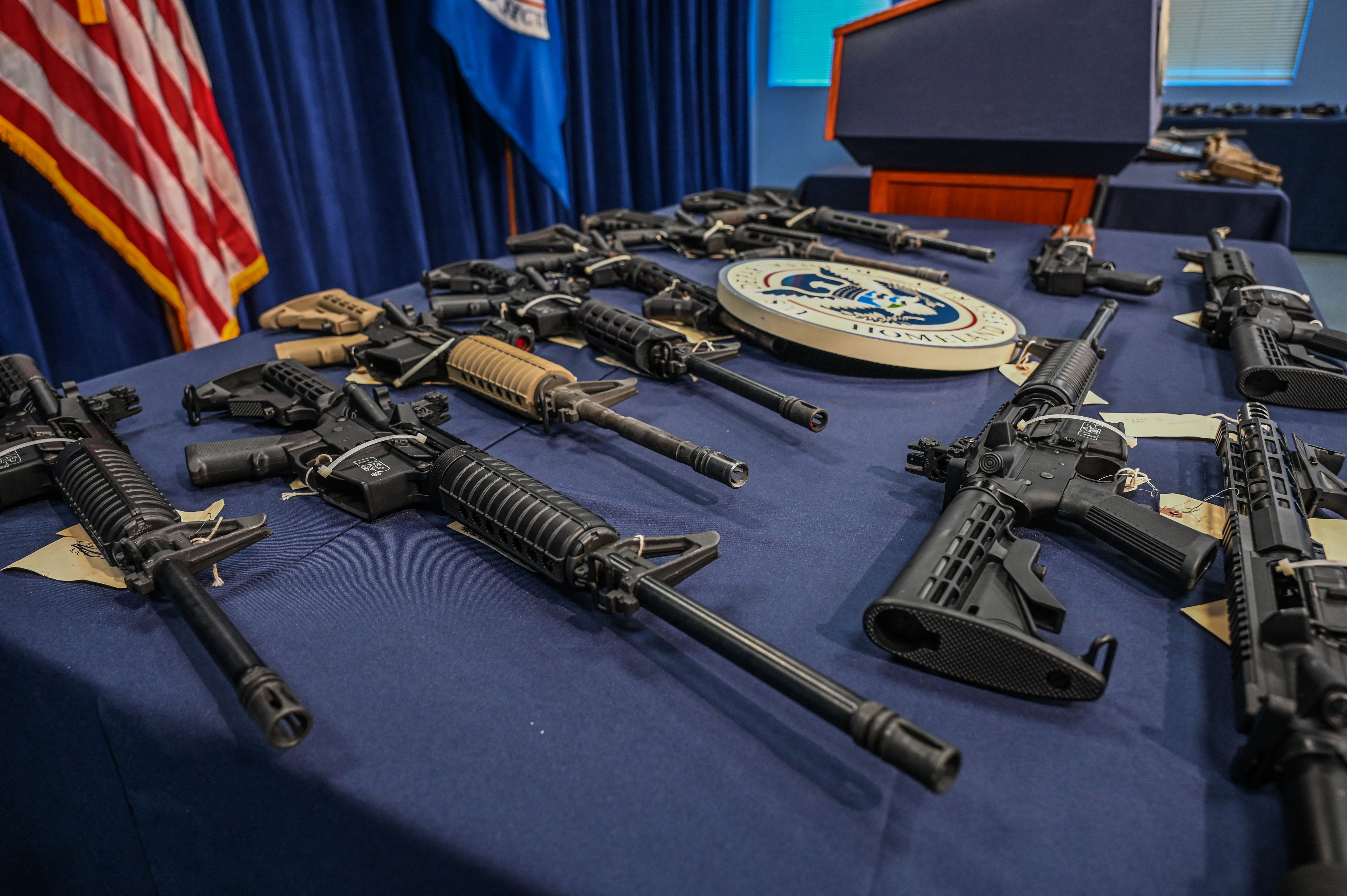 A collection of rifles lays displayed on a blue tablecloth, with a Department of Homeland Security seal and podium in the background, alongside an American flag.