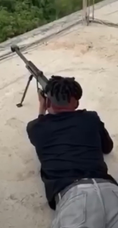 A man with dreadlocks is lying prone on a rooftop, firing a long-barreled rifle with a bipod toward the distance, with greenery visible in the background.