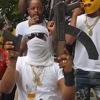 A group of individuals is gathered outdoors, with one prominently displaying firearms. They are wearing casual clothing and some have face coverings, sunglasses and jewelry.