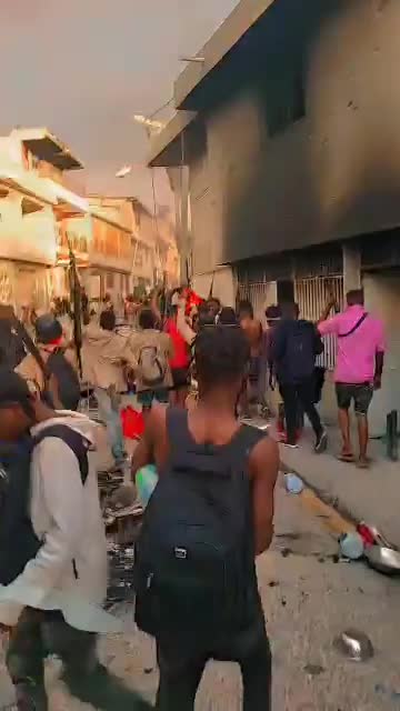 A group of people are barging down an urban street carrying rifles, near buildings with signs of fire damage.